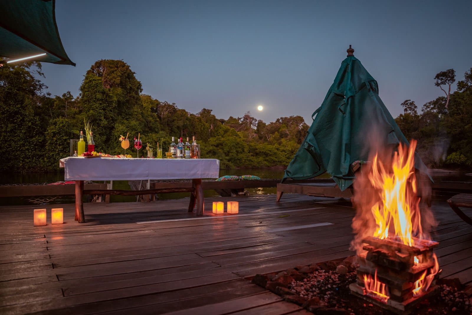 Cocktail hour hits differently when the moon shows up uninvited. An open timber deck at Cristalino Jungle Lodge at dusk, with a roaring bonfire in the foreground, paper lantern candles lining the floor, a white-clothed cocktail table set with colorful drinks and bottles, and a full moon rising above the rainforest treeline