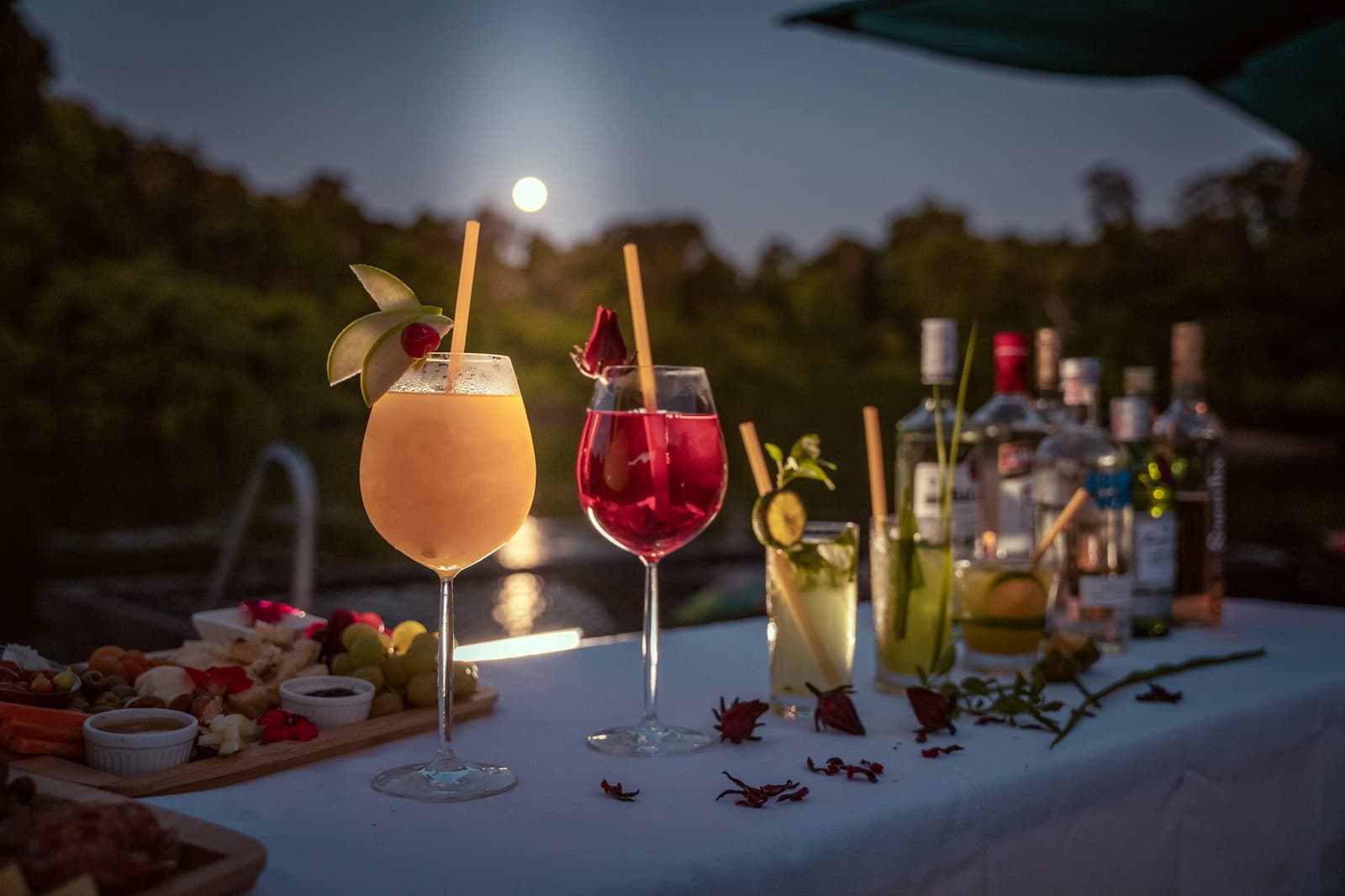 The full moon showed up right on time for happy hour. Four garnished cocktails and a charcuterie board on a white-draped table at Cristalino Jungle Lodge, with a full moon rising over the rainforest canopy in the background