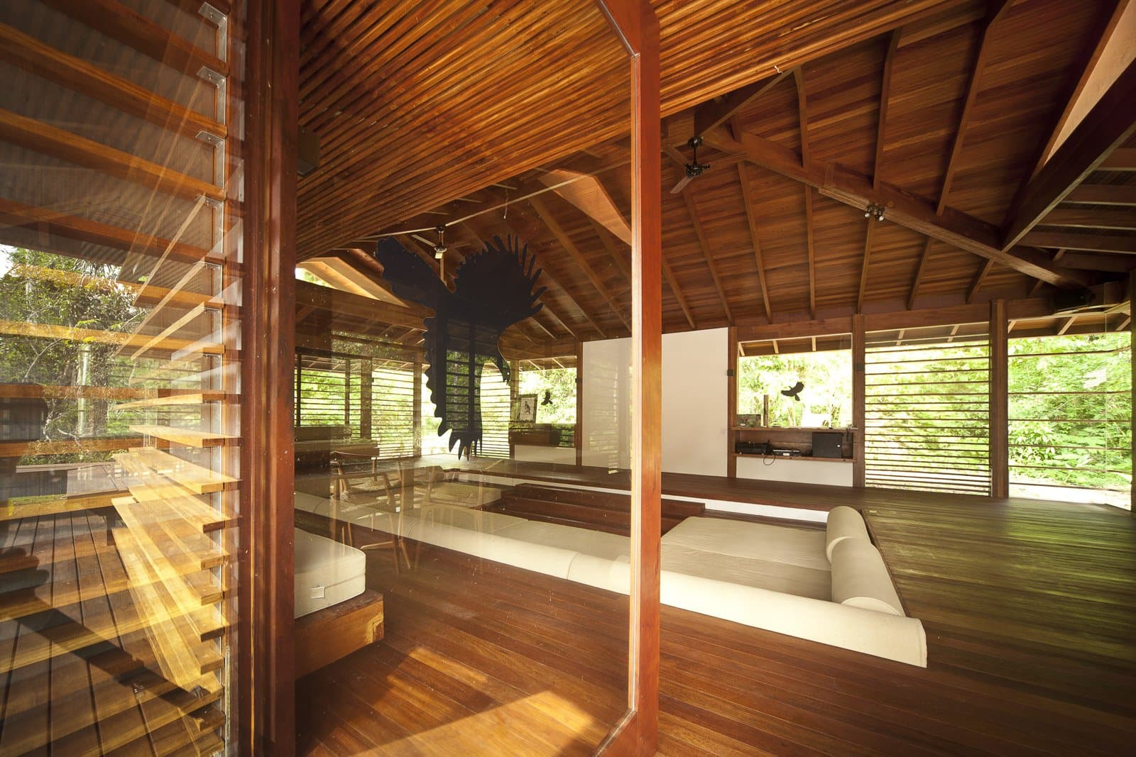 Hardwood, open walls, and the forest doing the decorating. Open-air common lounge at Cristalino Jungle Lodge featuring warm hardwood floors, a vaulted timber ceiling, white day-bed sofas, and louvred walls opening onto lush green rainforest