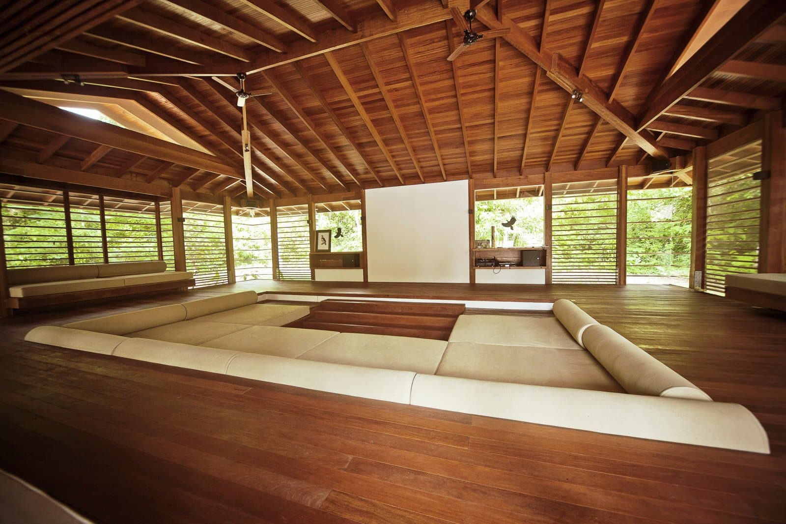 The kind of room that makes you forget your phone exists. The open-air lounge at Cristalino Jungle Lodge features a sunken seating area with cream cushions, polished hardwood floors, a large projector screen, and a vaulted timber roof with ceiling fans, surrounded by lush green forest on all sides.