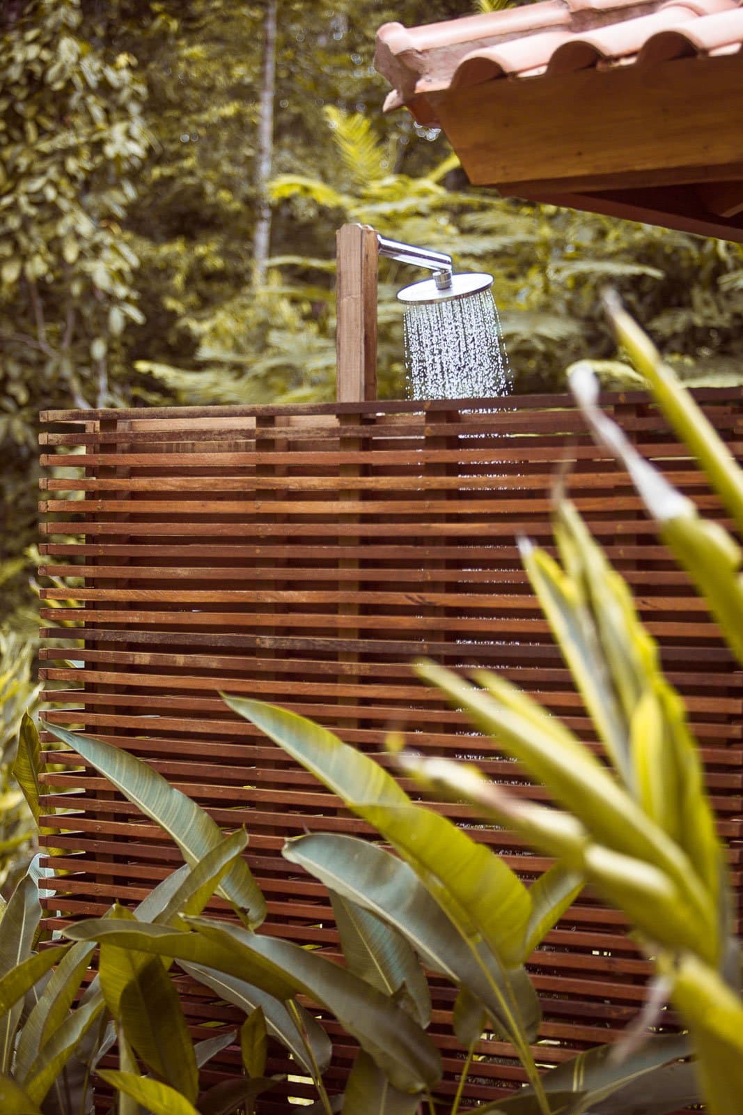 Rinsing off the day with the forest still close enough to touch. An open-air rain showerhead flowing with water above a slatted hardwood privacy screen at Cristalino Jungle Lodge, surrounded by lush tropical foliage and a terracotta-tiled roof