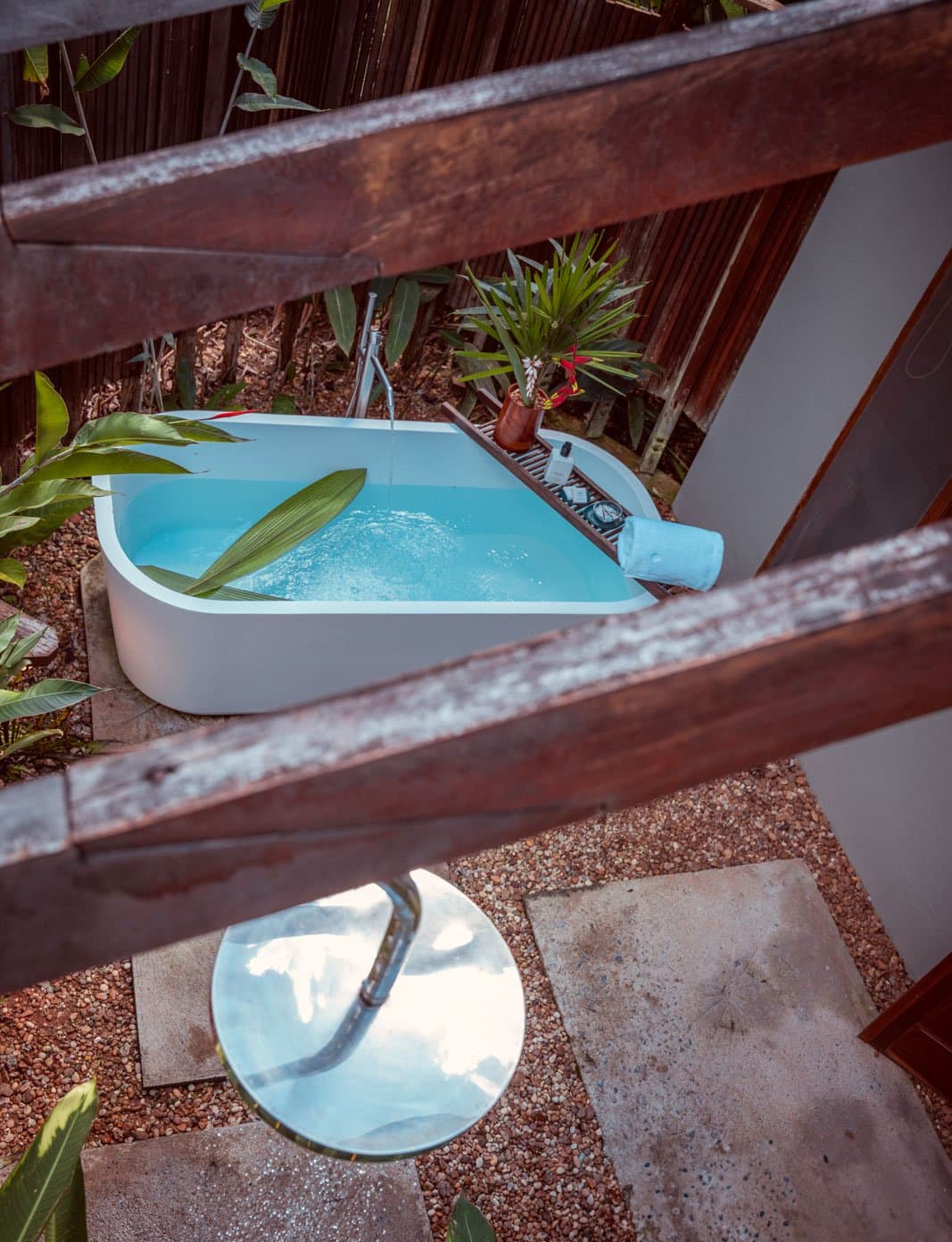 A private soak in the forest, with birds as your only company. An open-air freestanding soaking tub filled with clear blue water at Cristalino Jungle Lodge, surrounded by tropical foliage, a wooden bath caddy with flowers and toiletries, and a round outdoor shower below, viewed from above through a rustic timber railing