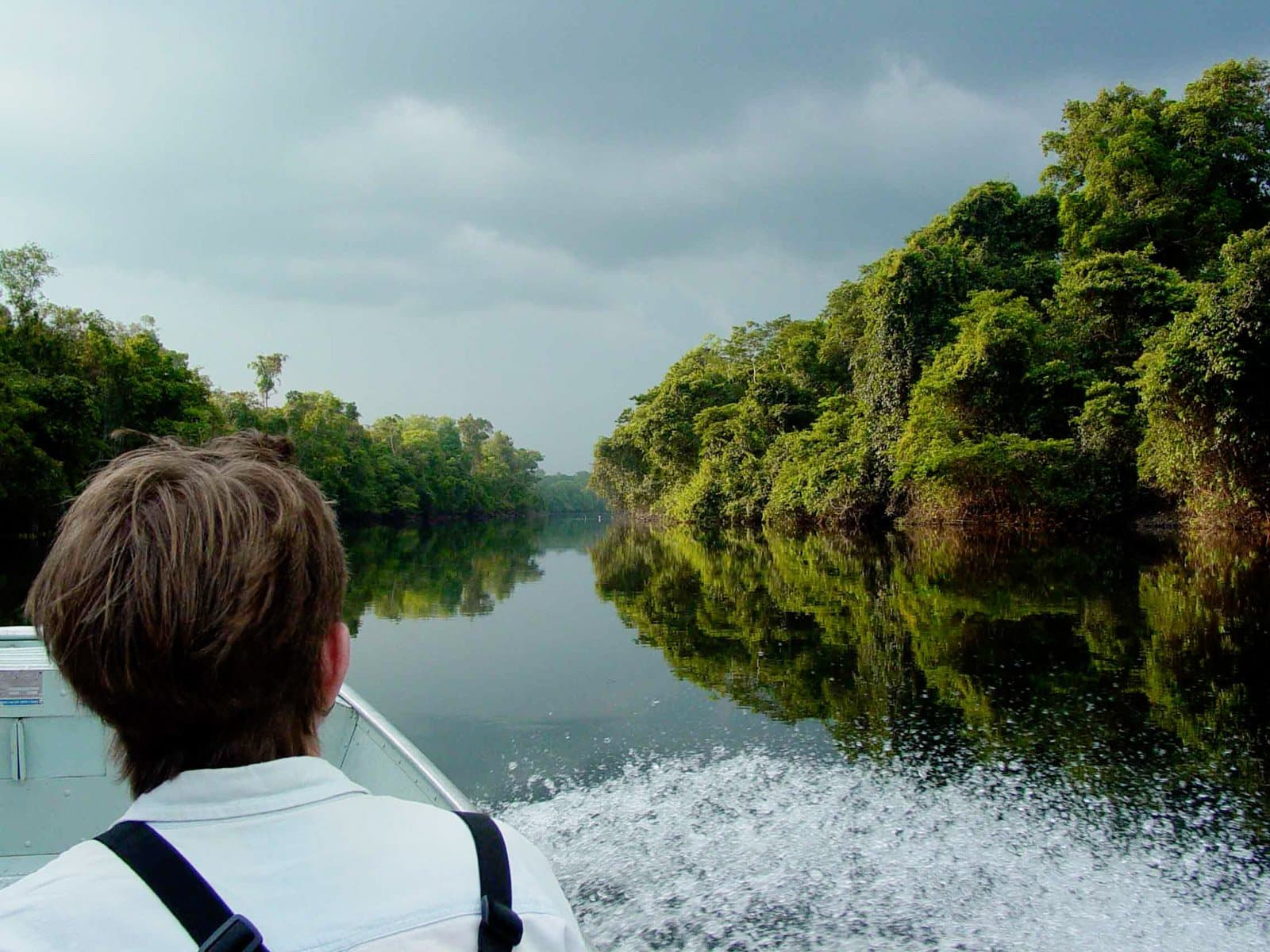 The river keeps its secrets until you're already moving. A passenger looks ahead from a motorboat gliding along a mirror-calm jungle river flanked by dense rainforest canopy at Cristalino Jungle Lodge