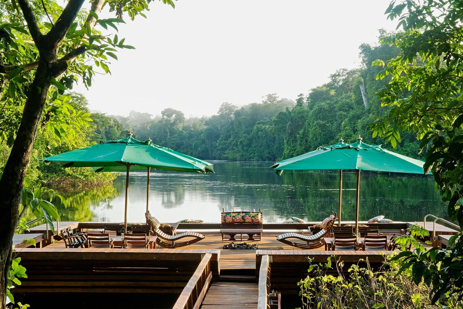 Your afternoon plans, sorted: a river view and nowhere to be. A wooden deck terrace at Cristalino Jungle Lodge with teak sun loungers and green parasols overlooking a calm jungle river flanked by dense rainforest