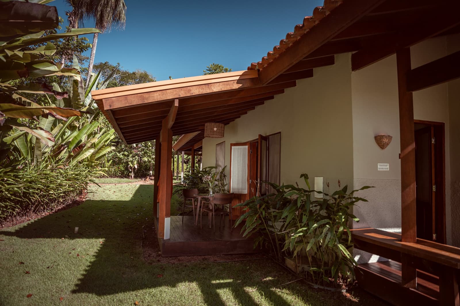 Your own patch of green, just steps from the forest. A timber-framed covered veranda at Cristalino Jungle Lodge, with a small outdoor dining table, rattan pendant lamp, and lush tropical garden stretching beyond under a clear blue sky