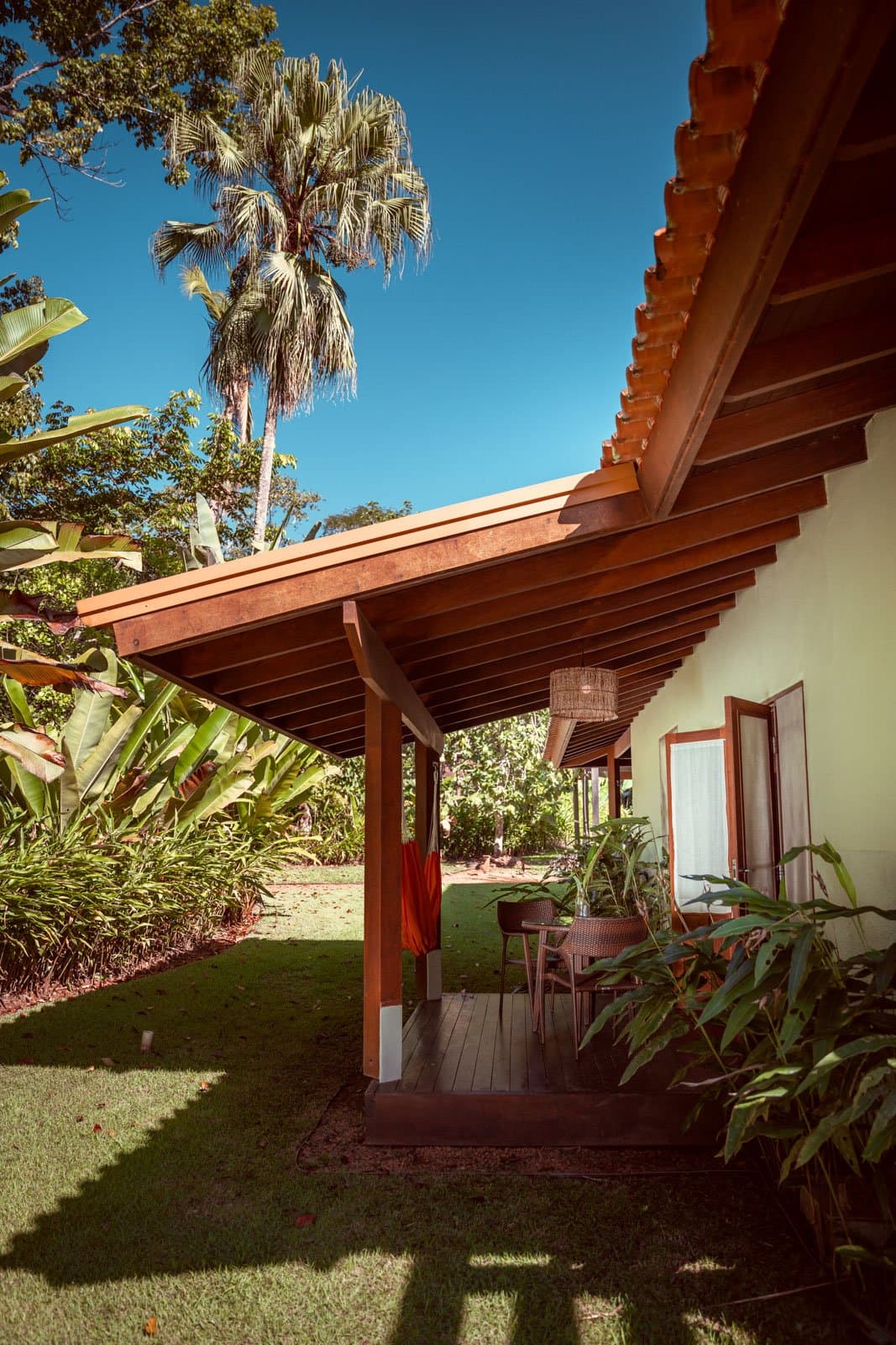 Shade, a rattan lamp, and the Amazon just beyond the lawn. A shaded hardwood veranda at Cristalino Jungle Lodge with a rattan pendant lamp, rattan chairs, and lush tropical garden including banana plants and a tall palm under a deep blue sky
