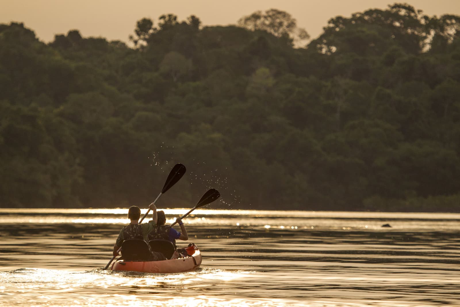The river keeps its own pace, and for once, so do you. Two paddlers in a tandem kayak glide across a glassy river at golden hour, with dense rainforest canopy reflected on the water at Cristalino Jungle Lodge