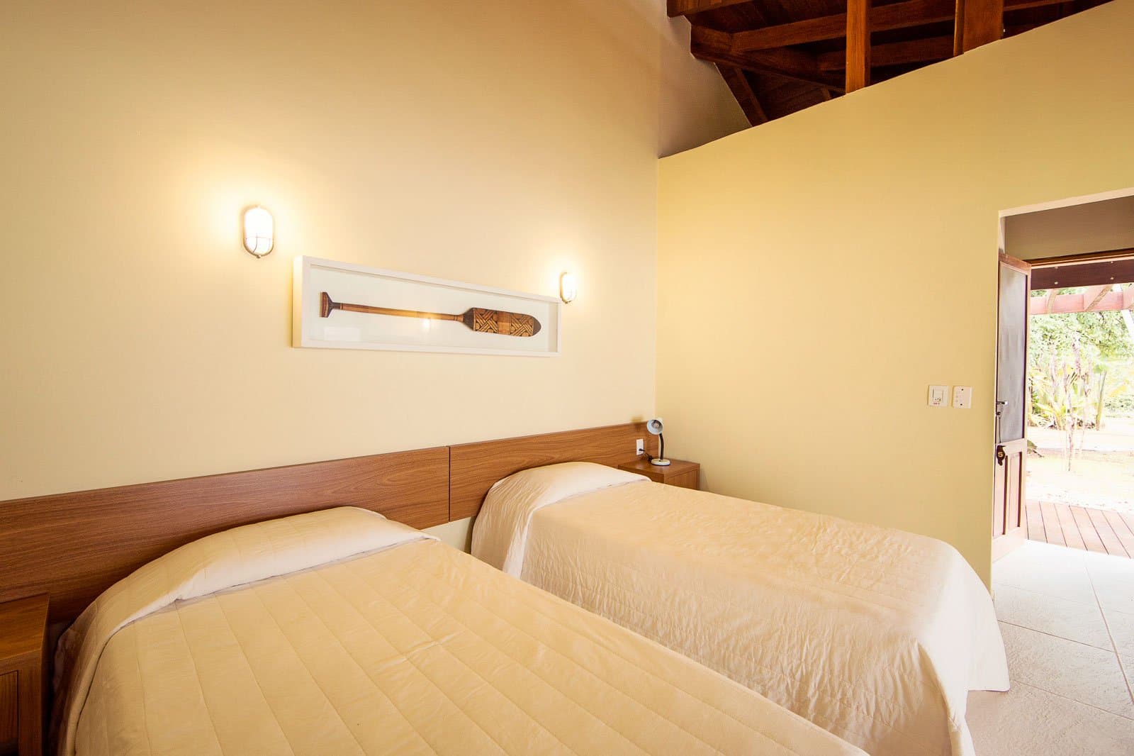 Cosy, clean, and a wooden paddle on the wall, the Amazon has its own kind of décor. Twin-bed room at Cristalino Jungle Lodge with warm timber headboard, cream quilted bedding, a framed decorative paddle on the yellow wall, exposed wooden rafters, and an open door leading to a lush garden terrace