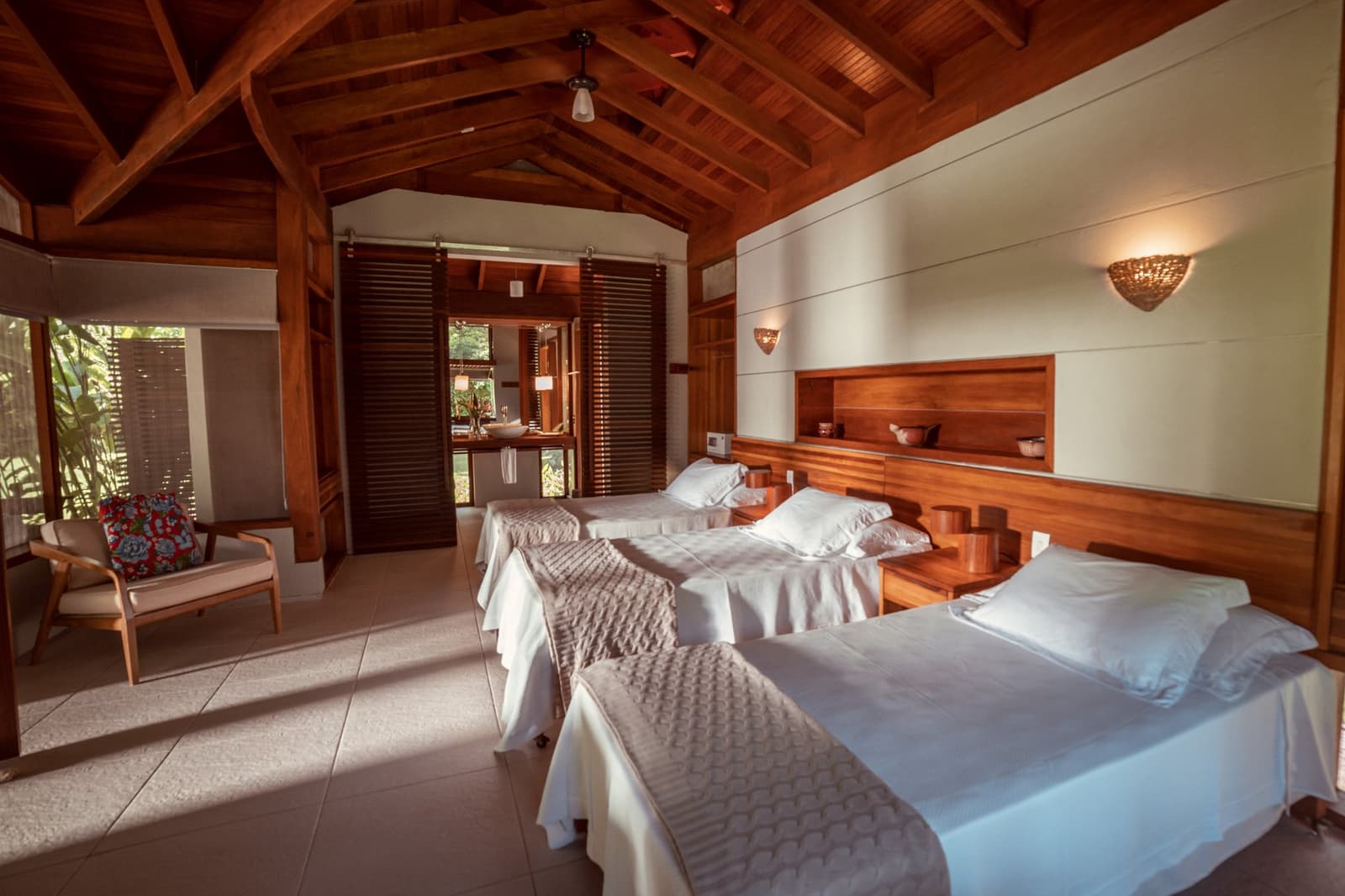 Solid wood, white linen, and the jungle just outside the door. Twin-bed room at Cristalino Jungle Lodge with vaulted timber ceiling, warm wall sconces, a wooden headboard, and tropical garden views through louvred doors