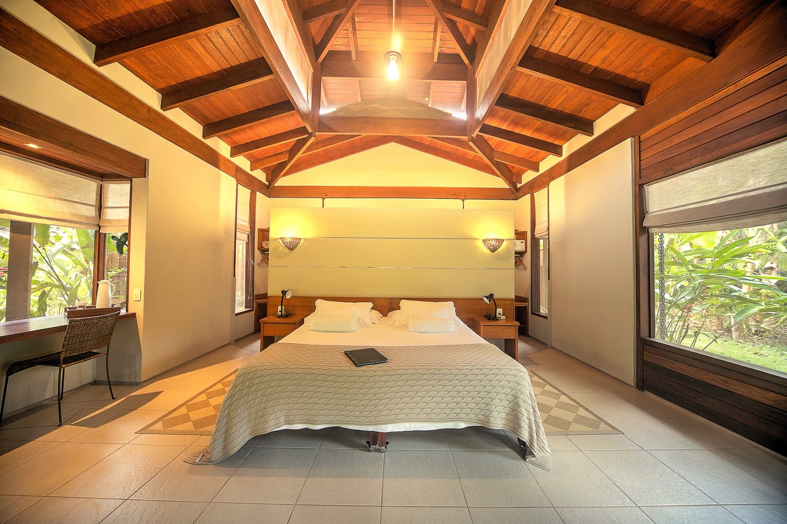 Tall ceilings, green views on every side, and the forest just a step outside your window. A spacious bedroom at Cristalino Jungle Lodge featuring a vaulted hardwood-beam ceiling, a king-size bed with white linen, warm wall sconces, and lush tropical greenery visible through wide windows on both sides