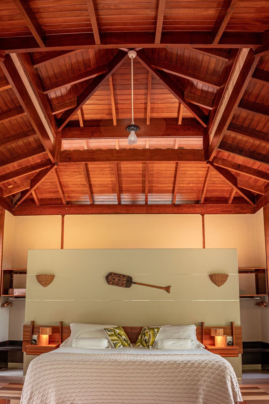 High ceilings, warm wood, and a paddle on the wall, this room knows where it is. A soaring vaulted hardwood ceiling rises above a double bed at Cristalino Jungle Lodge, flanked by glowing wooden nightstands and a decorative paddle mounted on the headboard wall