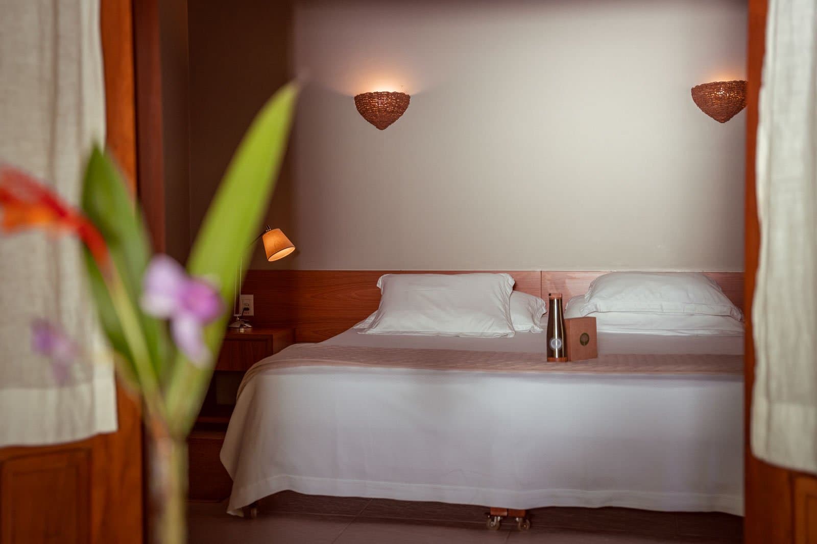 A good night's sleep in the Amazon is easier than you'd think. A neatly made double bed with crisp white linen and warm timber wall sconces in a Cristalino Jungle Lodge guest room, framed by a blurred tropical flower in the foreground