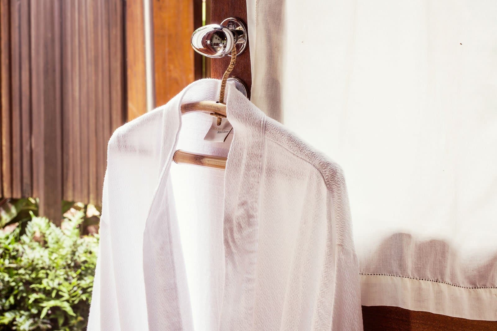 Comfort waiting right where you left it. A plush white terrycloth robe hanging on a bamboo hanger from a polished door hook at Cristalino Jungle Lodge, with tropical greenery visible through the window behind
