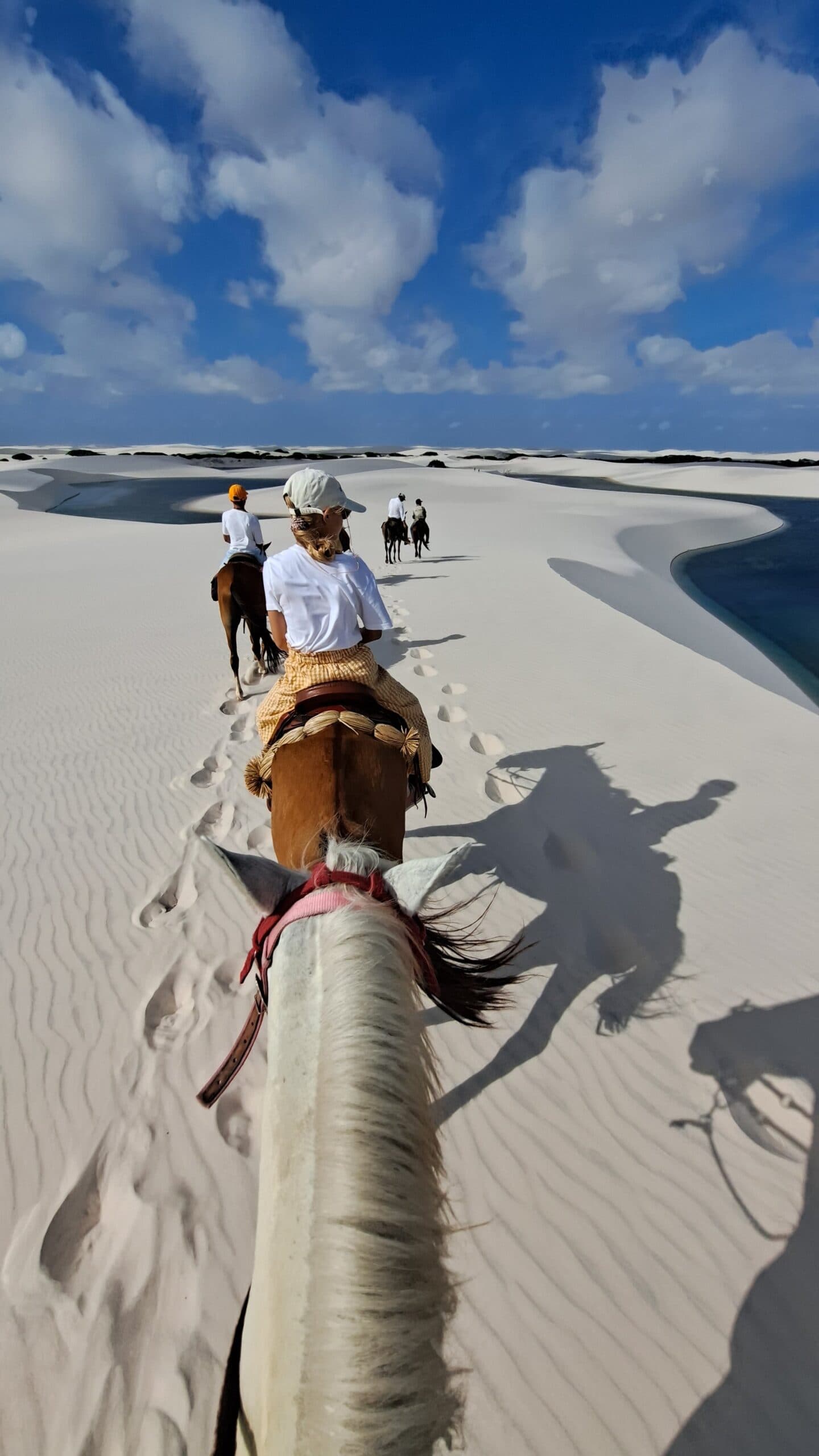 Hoofprints in white sand, a lagoon just off the trail. Not a bad commute. Point-of-view shot from horseback on a horseback riding Lençóis Maranhenses tour, with riders and hoofprints tracing a line across rippled white dunes beside a turquoise lagoon under a bright blue sky