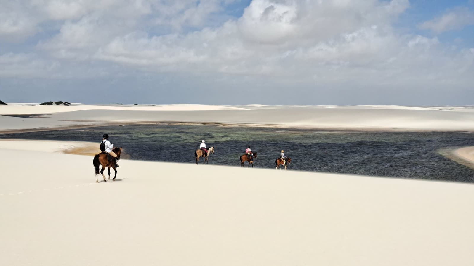 The only tracks out here belong to horses. Four riders on horseback cross white sand dunes beside a dark freshwater lagoon on a horseback riding Lençóis Maranhenses tour, under a partly cloudy sky
