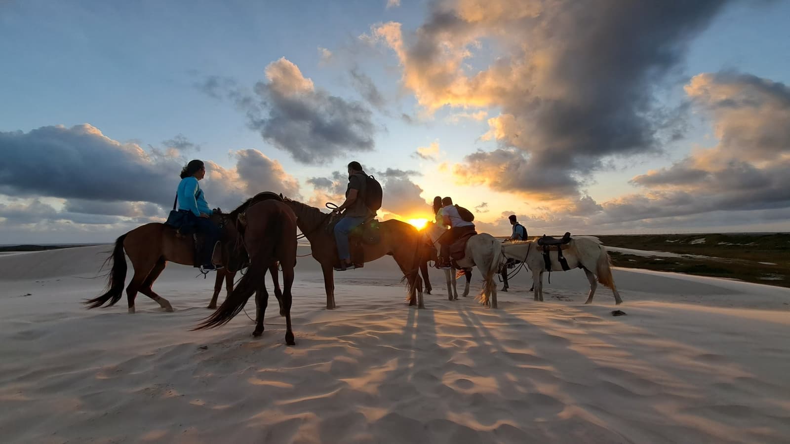The dunes have a way of making every hour feel like the right one. A group of riders on horseback silhouetted against a blazing sunset over the white sand dunes of Lençóis Maranhenses, with dramatic clouds spreading across the sky during a dune crossing on horseback