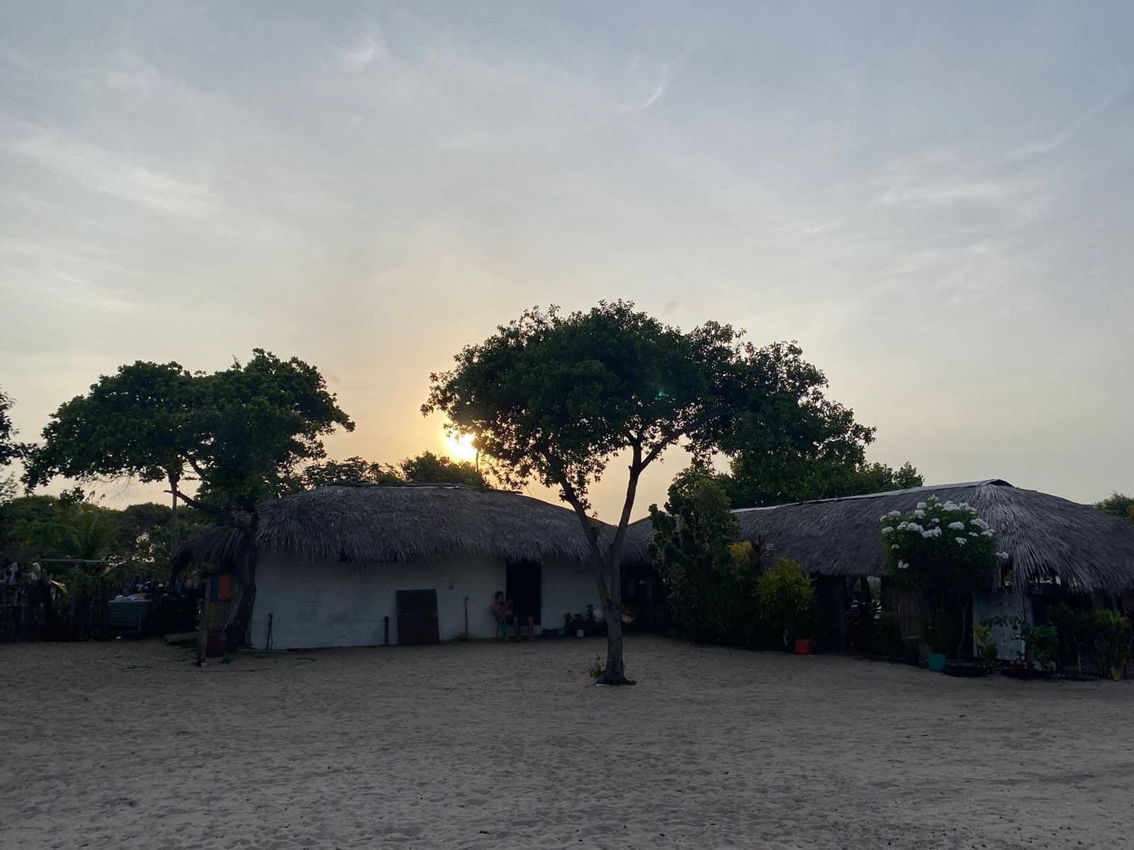 The kind of place where the day winds down slowly and nobody rushes dinner. Whitewashed thatched-roof houses and a lone tree silhouetted against the setting sun on a sandy open ground, a typical local community along the horseback riding Lençóis Maranhenses tour route