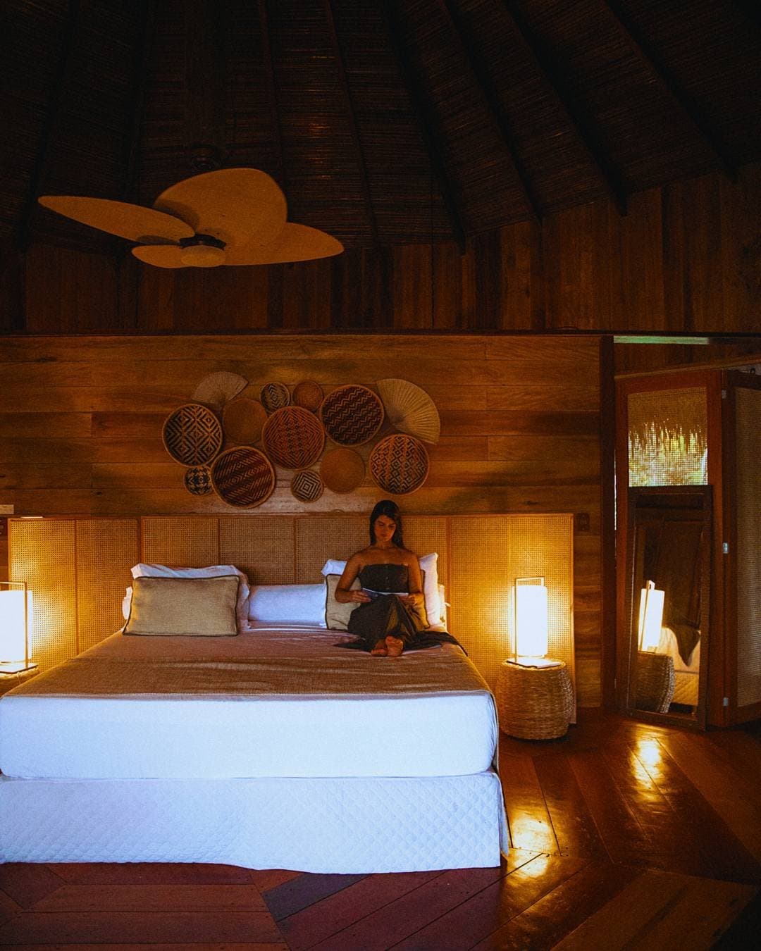 Some evenings, a good book and warm lamplight are exactly enough. A woman reads on a king-size bed inside a Juma Amazon Jungle Lodge bungalow, surrounded by warm lamplight, a woven rattan headboard, decorative woven baskets on timber walls, and a thatched ceiling with a wooden fan