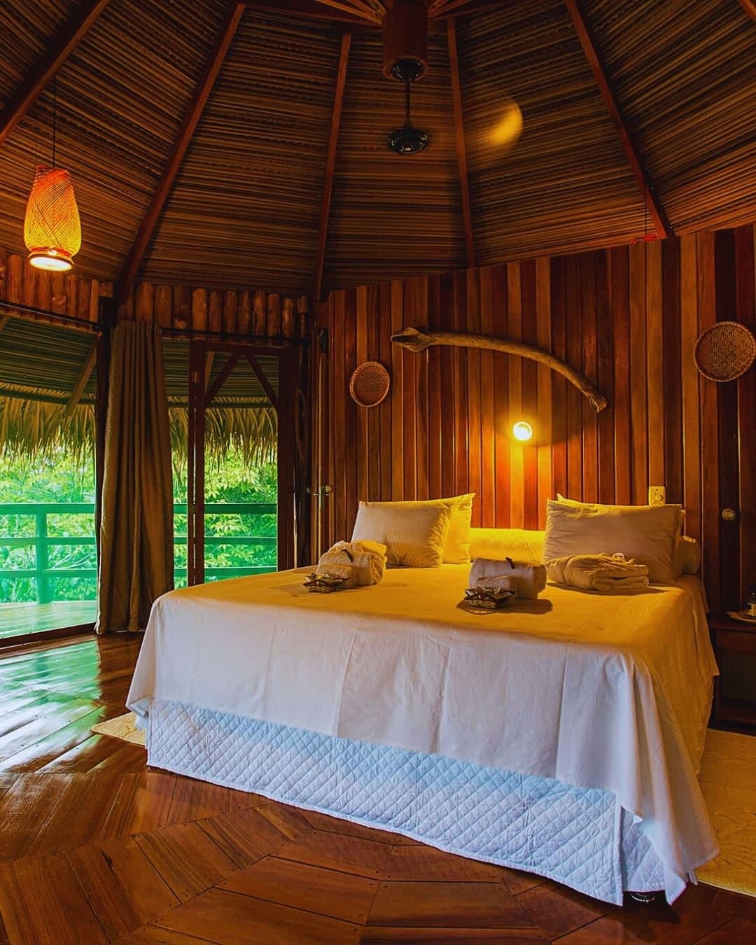 Fall asleep to the forest, wake up still inside it. Warm-lit bungalow interior at Juma Amazon Jungle Lodge featuring a double bed with white linen, thatched conical ceiling, polished hardwood floor, woven pendant lamp, and an open balcony door framing lush rainforest vegetation
