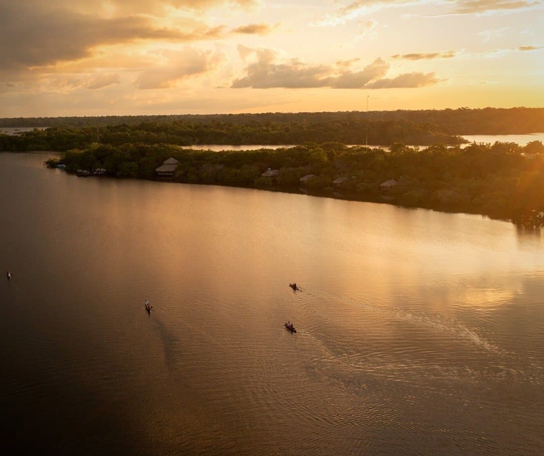 The Amazon has its own clock, and golden hour is non-negotiable. Aerial view of small canoes gliding across a wide river at golden sunset, with the thatched bungalows of Juma Amazon Jungle Lodge nestled among dense rainforest on the far bank