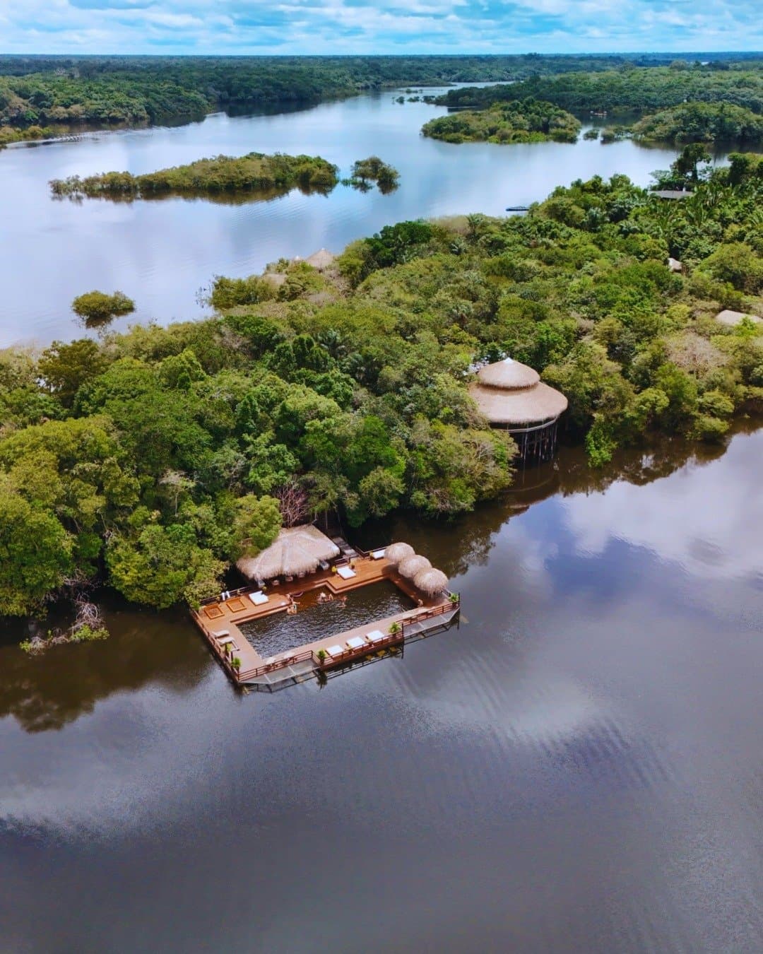 Your own slice of river, tucked into the canopy. Aerial view of Juma Amazon Jungle Lodge showing a wooden floating deck with thatched-roof pavilions and a river pool, surrounded by flooded rainforest and dark Amazonian waters
