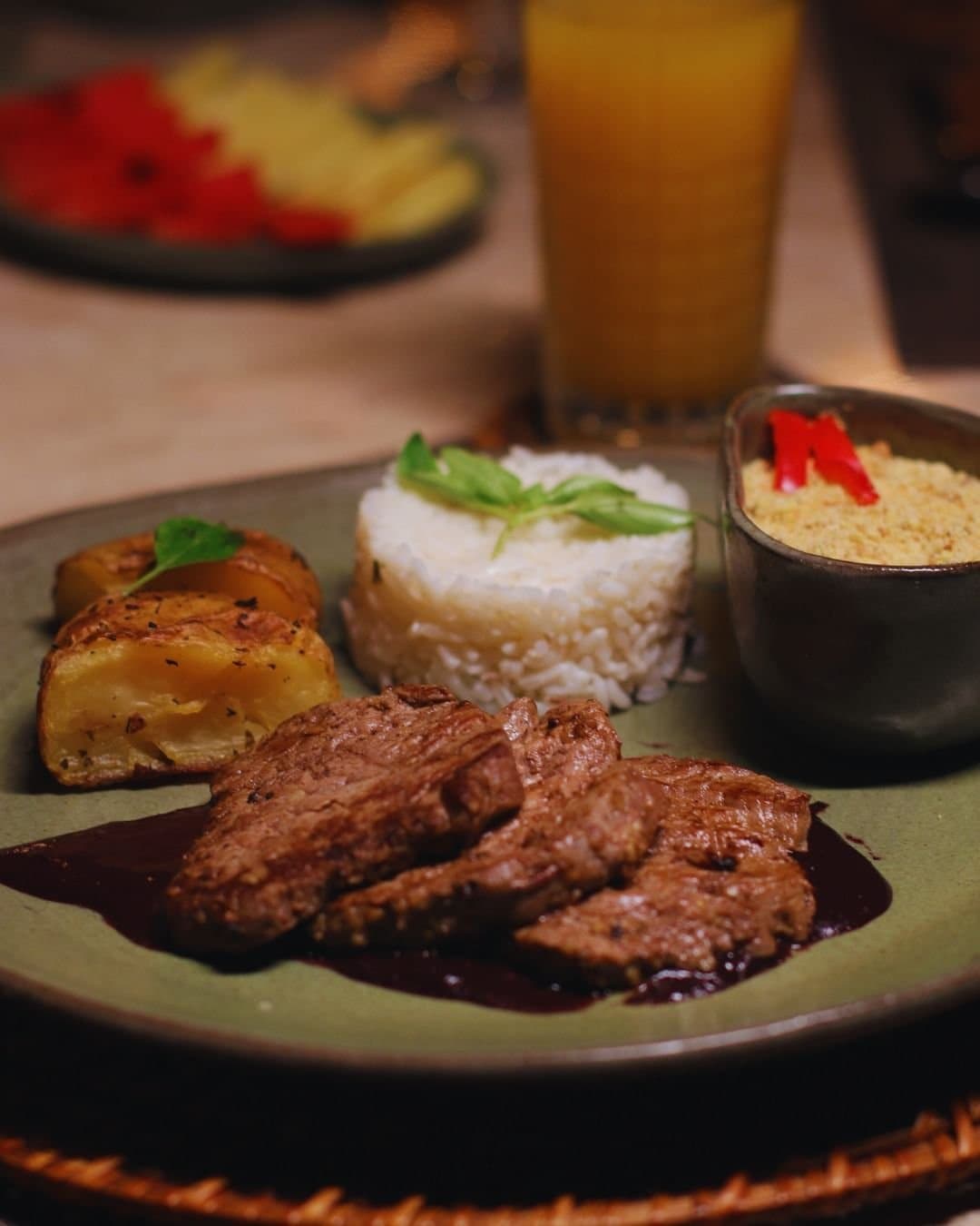 Lodge cooking that takes Brazilian comfort food seriously. A plated dinner at Juma Amazon Jungle Lodge featuring sliced grilled beef in dark sauce, a moulded rice garnished with fresh herbs, herb-roasted potato wedges, and a small bowl of farofa topped with red pepper strips, served alongside a glass of orange juice