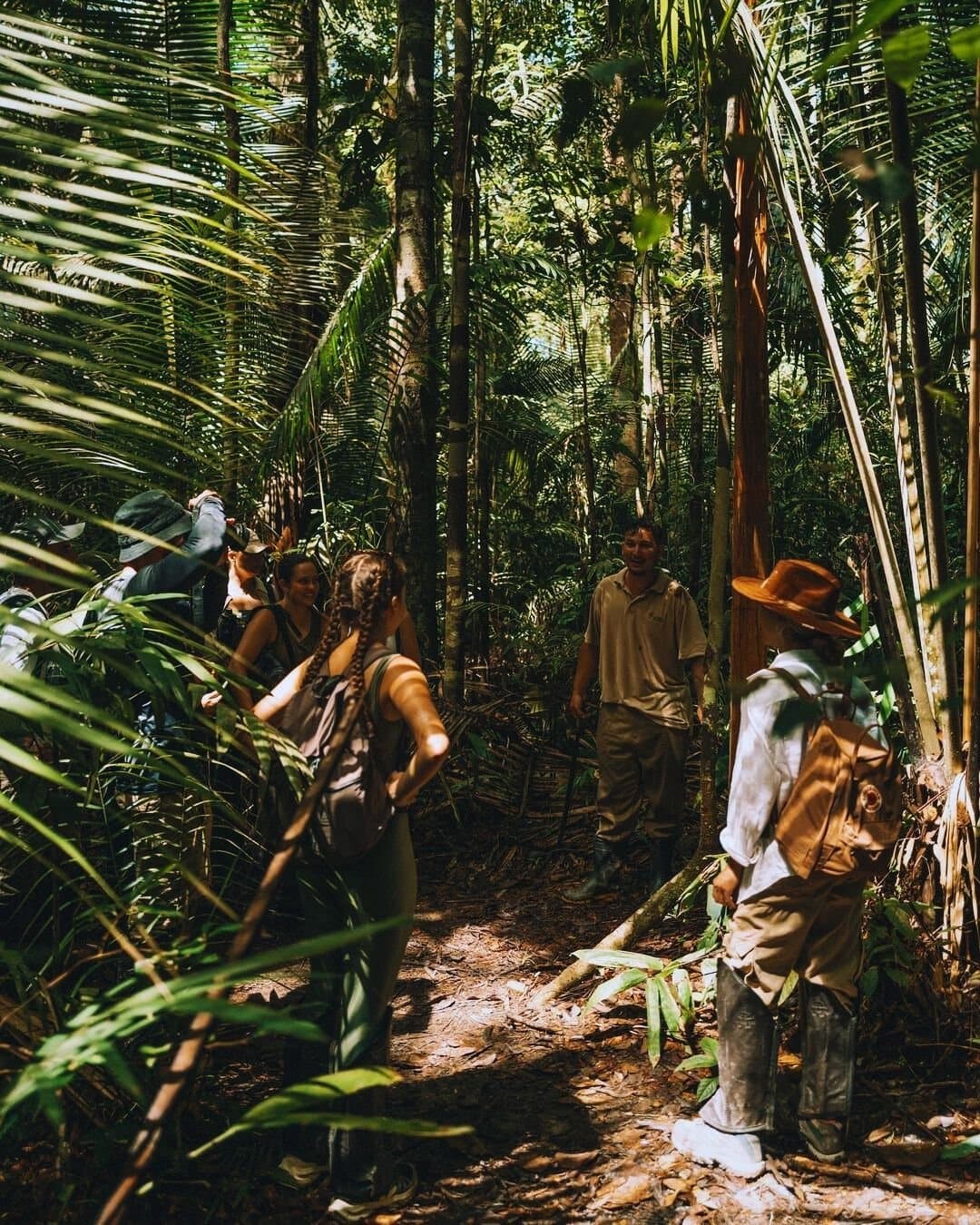 The forest has its own rhythm, and a good guide helps you hear it. A small group of smiling travellers follows a local guide along a narrow trail through dense tropical rainforest at Juma Amazon Jungle Lodge, with palm fronds and tall trees surrounding the path
