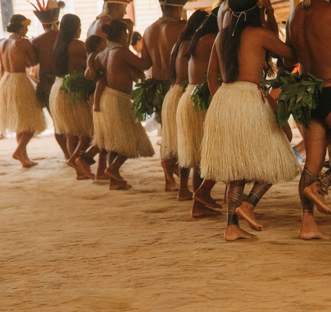Every step lands with intention. This is living culture, not a performance for cameras. A line of indigenous dancers wearing woven straw skirts and fresh leaf adornments move barefoot across a sand floor during a traditional ceremony at the Juma Amazon Jungle Lodge community visit
