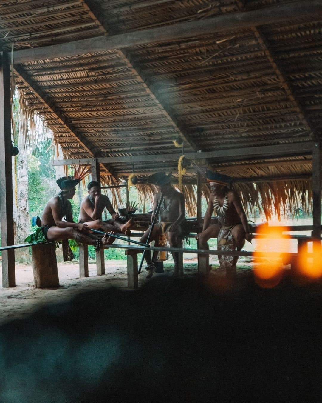 Life inside the maloca moves at its own pace, and you're welcome to slow down with it. Indigenous community members in traditional body paint, headdresses and beaded necklaces gathered under a thatched-roof open-air structure at Juma Amazon Jungle Lodge, long blowpipes resting across wooden benches