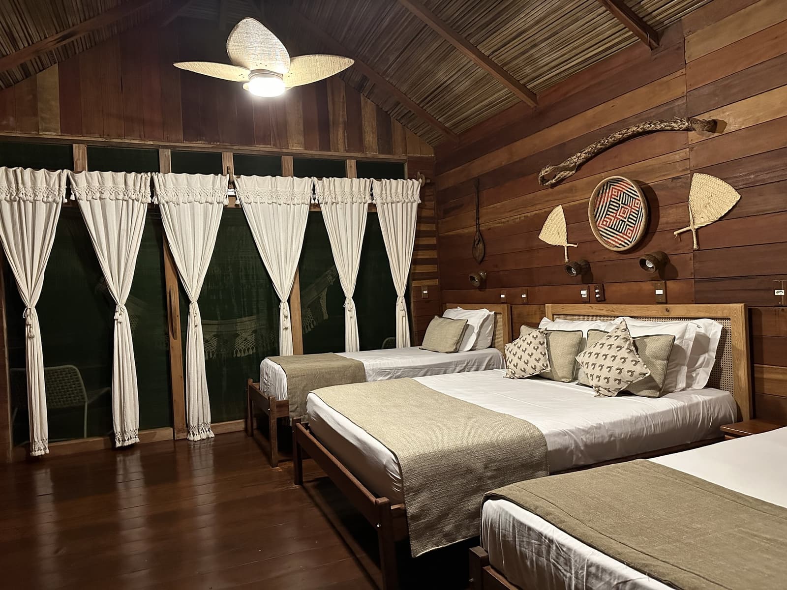 Rustic craft on the walls, crisp linen on the beds, comfort that actually fits the forest. A spacious Juma Amazon Jungle Lodge bungalow at night featuring three wooden-framed beds dressed in white linens, a woven-blade ceiling fan, floor-to-ceiling draped windows, and indigenous woven baskets and paddles decorating the timber walls