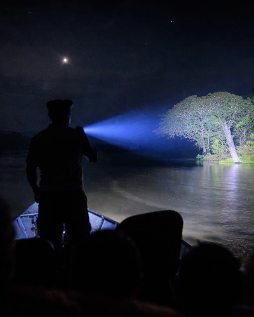 Out here at night, the river belongs to whoever holds the torch. A silhouetted guide standing on the bow of a small boat shines a powerful spotlight across a dark river toward a large tree at night during a Juma Amazon Jungle Lodge night tour, with a crescent moon visible above