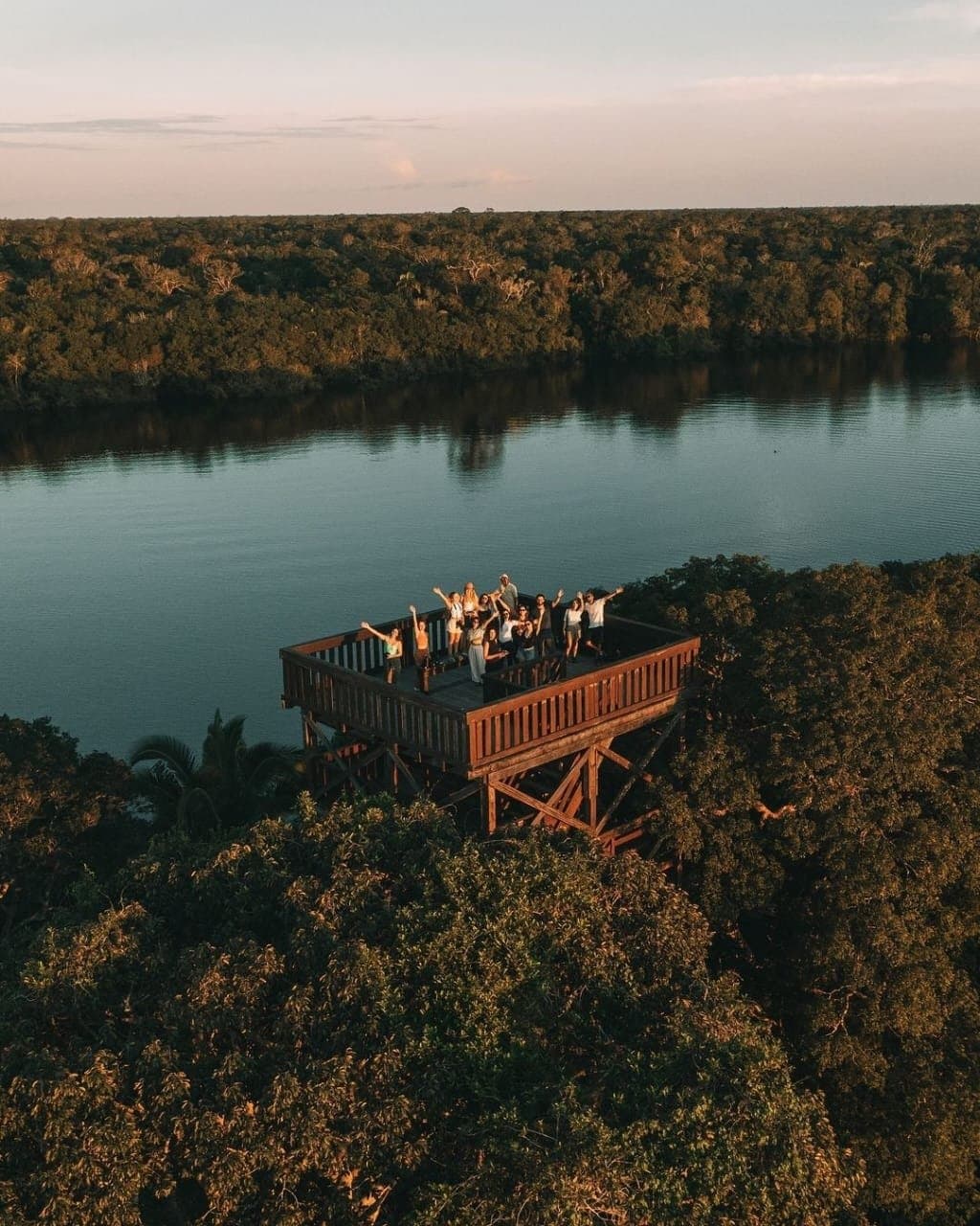 Arms up, canopy below, river ahead, this is how the Amazon ends a day. A group of excited travellers waving from a tall wooden observation tower at Juma Amazon Jungle Lodge, surrounded by dense rainforest canopy and a calm river at sunset