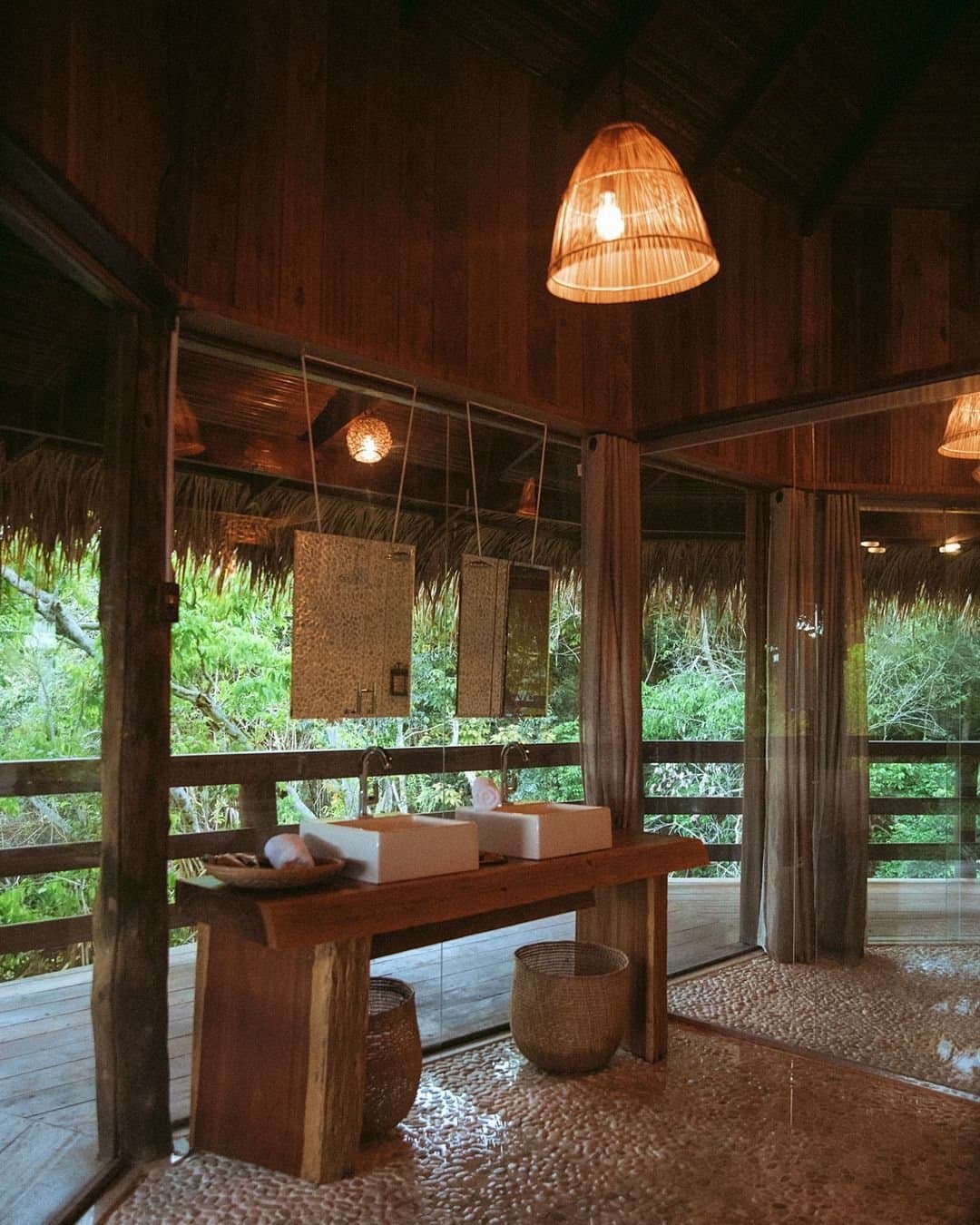 Showering with the forest watching, no complaints from either side. Open-air bathroom at Juma Amazon Jungle Lodge with twin vessel sinks on a raw-timber vanity, woven pendant lamp, pebble-stone floor, and floor-to-ceiling glass walls overlooking dense rainforest canopy