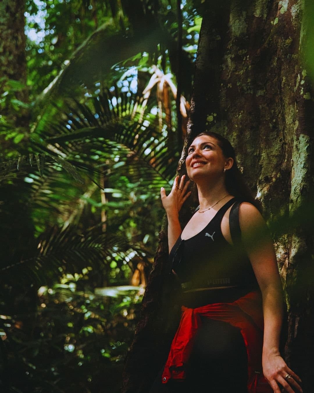 Some trees make you stop and look up. Every single time. A smiling woman leans against a massive rainforest tree trunk on a Juma Amazon Jungle Lodge guided hike, surrounded by lush ferns and towering palms
