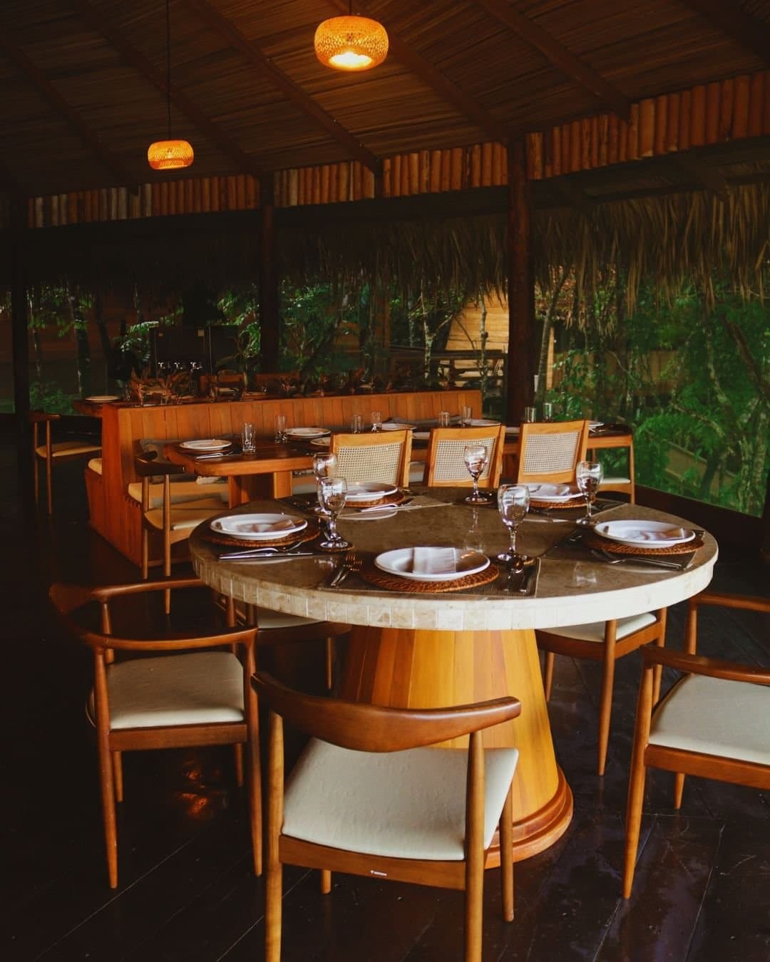 Dinner here feels like the jungle pulled up a chair and joined you. A set oval dining table with woven placemats, wine glasses and white plates inside the open-sided Juma Amazon Jungle Lodge restaurant, surrounded by bamboo walls, a thatched roof and lush rainforest greenery beyond