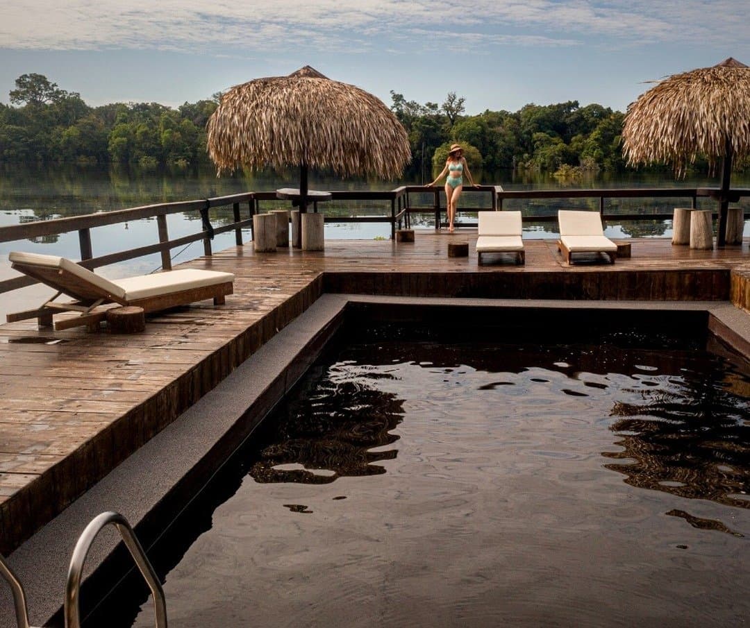 Dark-water pool, river view, zero obligations, the Amazon has its own pace. A dark-water river pool on a timber deck at Juma Amazon Jungle Lodge, with thatched palapa umbrellas, sun loungers, and a guest overlooking a calm jungle river