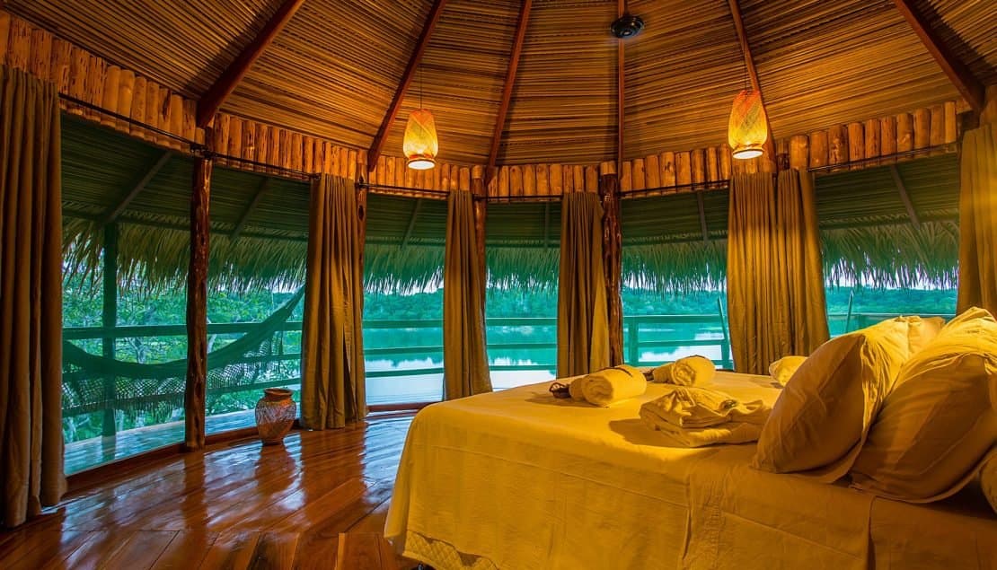 Your bedroom view is a river. The hammock outside is already calling. Inside a Juma Amazon Jungle Lodge bungalow with a king bed, woven pendant lamps, thatched ceiling, and panoramic open windows overlooking a calm jungle river