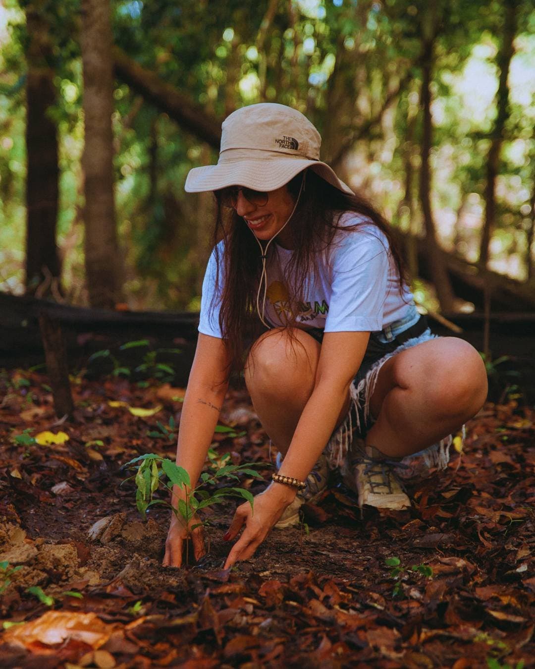 One seedling at a time, the forest gets a little help. A smiling woman in a wide-brim hat crouches to plant a young seedling in the forest floor at Juma Amazon Jungle Lodge, hands pressing dark soil around its roots
