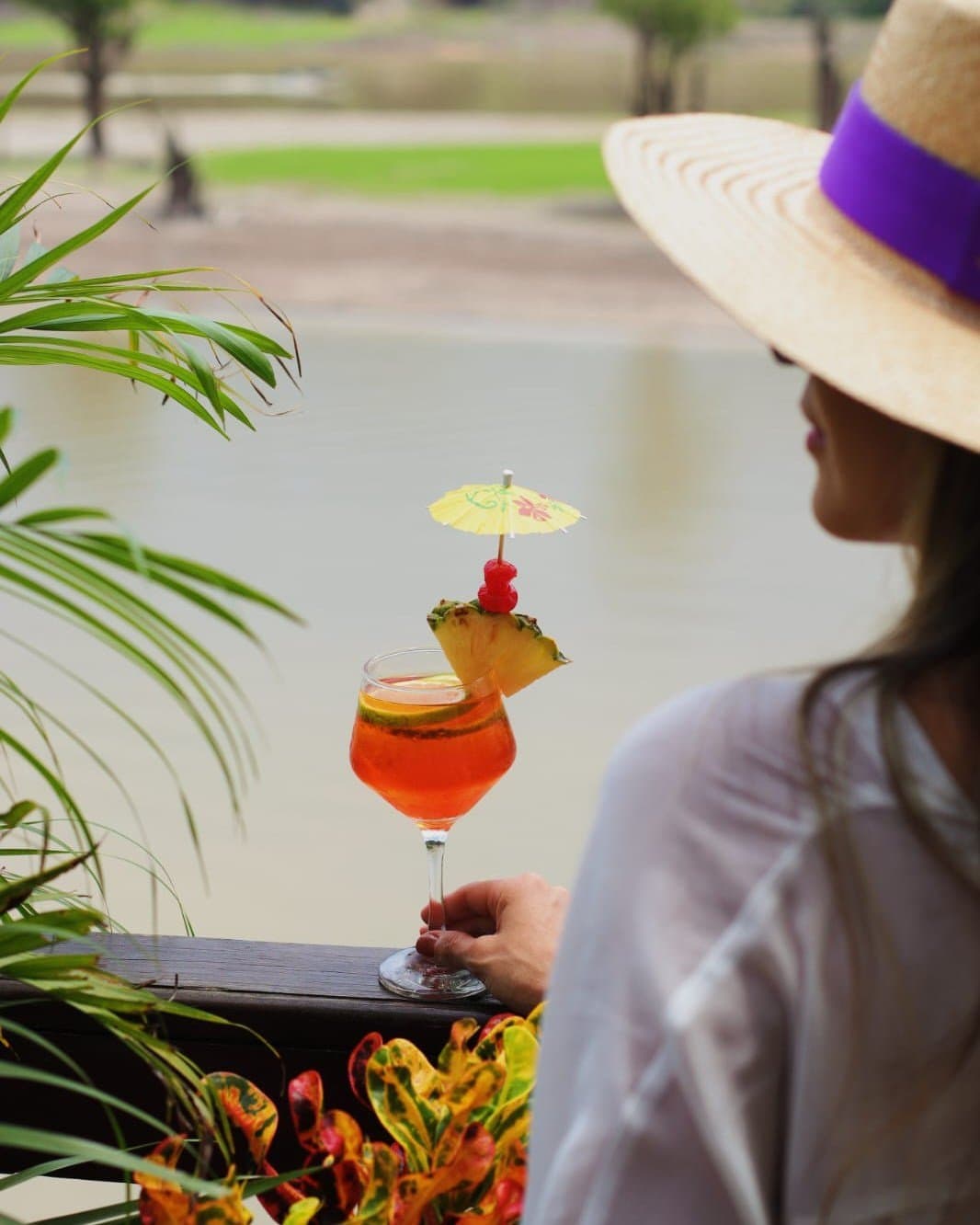 The cocktail hour hits differently when the Amazon is your backdrop. A woman in a wide-brimmed straw hat holds a vibrant orange tropical cocktail garnished with pineapple, cherry and a paper umbrella at Juma Amazon Jungle Lodge, with a tranquil waterway and lush greenery behind her