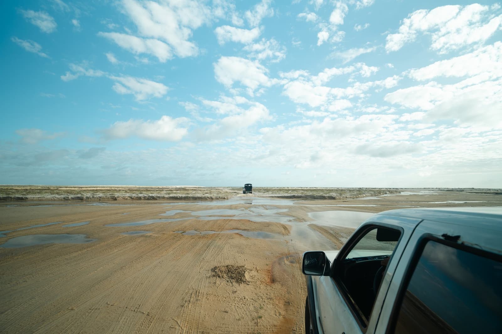 Out here, the road is whatever the tide left behind. Two 4x4 vehicles crossing a vast wet sandy flat under a wide blue sky on a Lençóis Maranhenses trip, with shallow puddles reflecting light across the trackless terrain