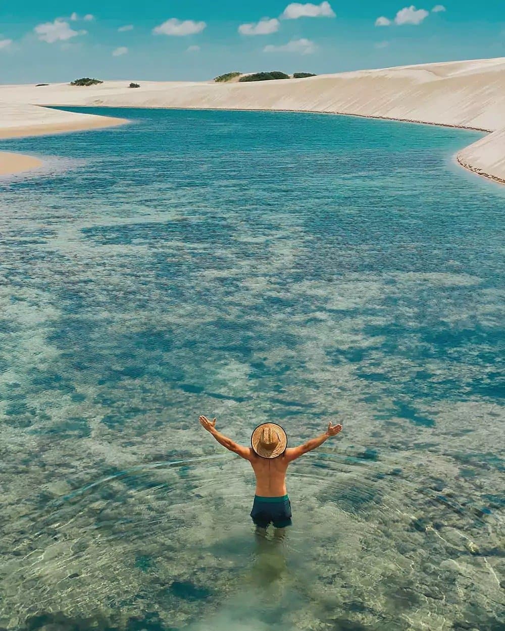 That first step into the lagoon hits different every single time. A traveler standing waist-deep in a crystal-clear turquoise lagoon on a Lençóis Maranhenses trip, arms raised wide, with sweeping white sand dunes stretching into the distance under a bright blue sky