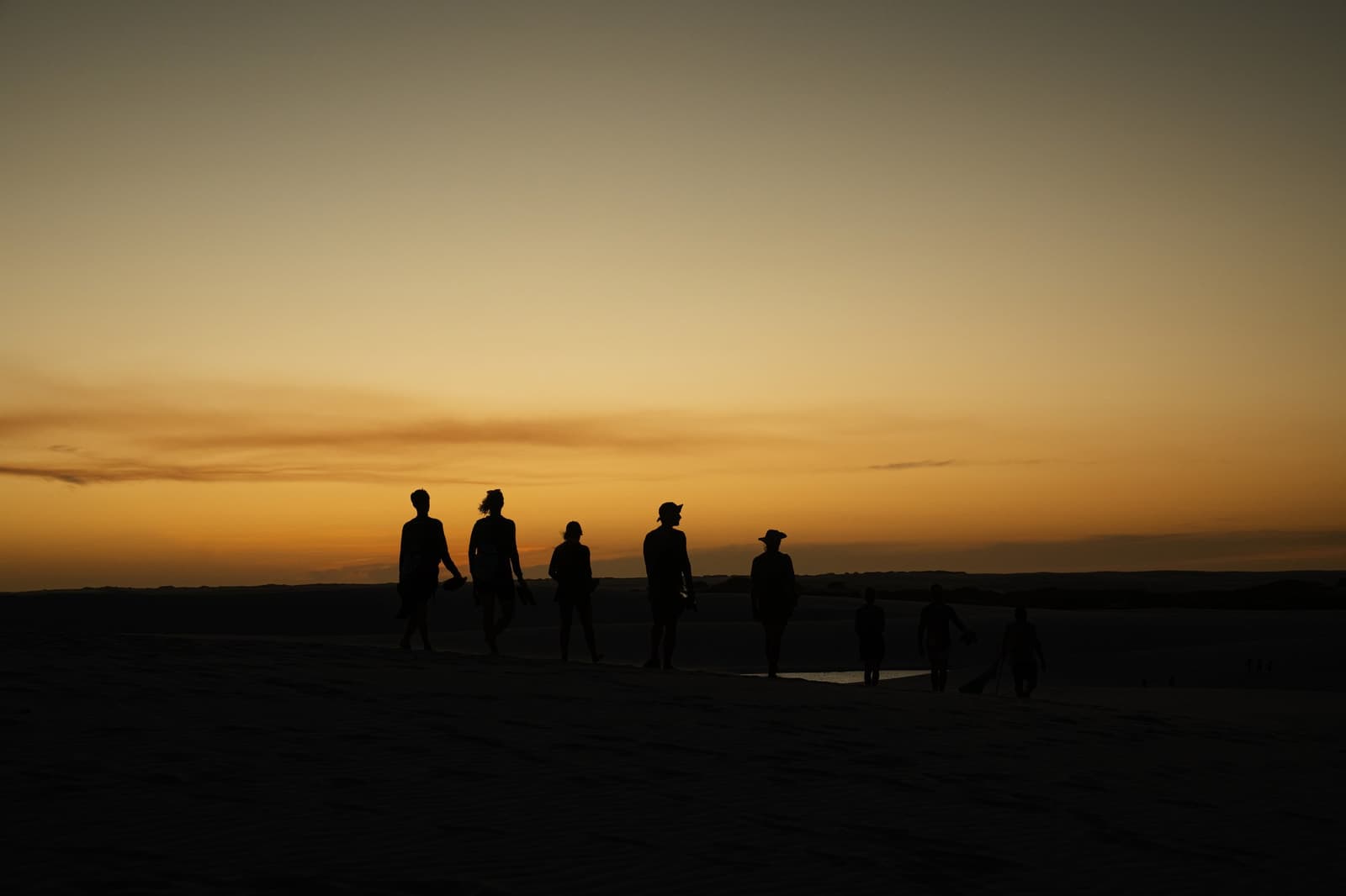 The dunes look different when the light decides to show off. Silhouettes of a group of travelers walking across open dunes during a Lençóis Maranhenses trip, set against a vivid amber and orange sunset sky