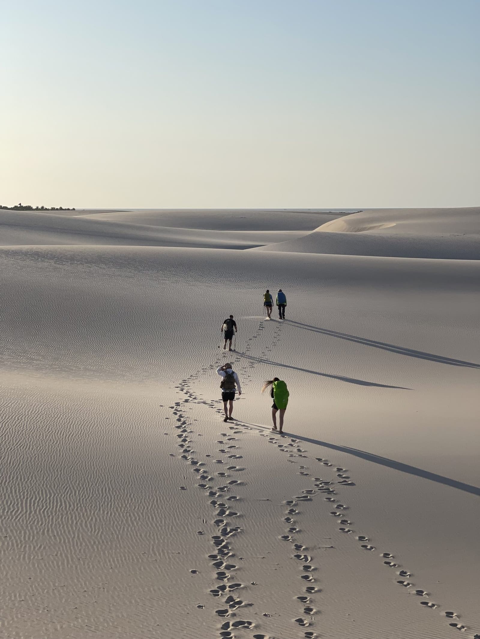 Four people, one trail, and a whole lot of sand ahead. Five hikers and their footprints crossing an expanse of rippled white sand dunes on a Lençóis Maranhenses trip, under a pale morning sky