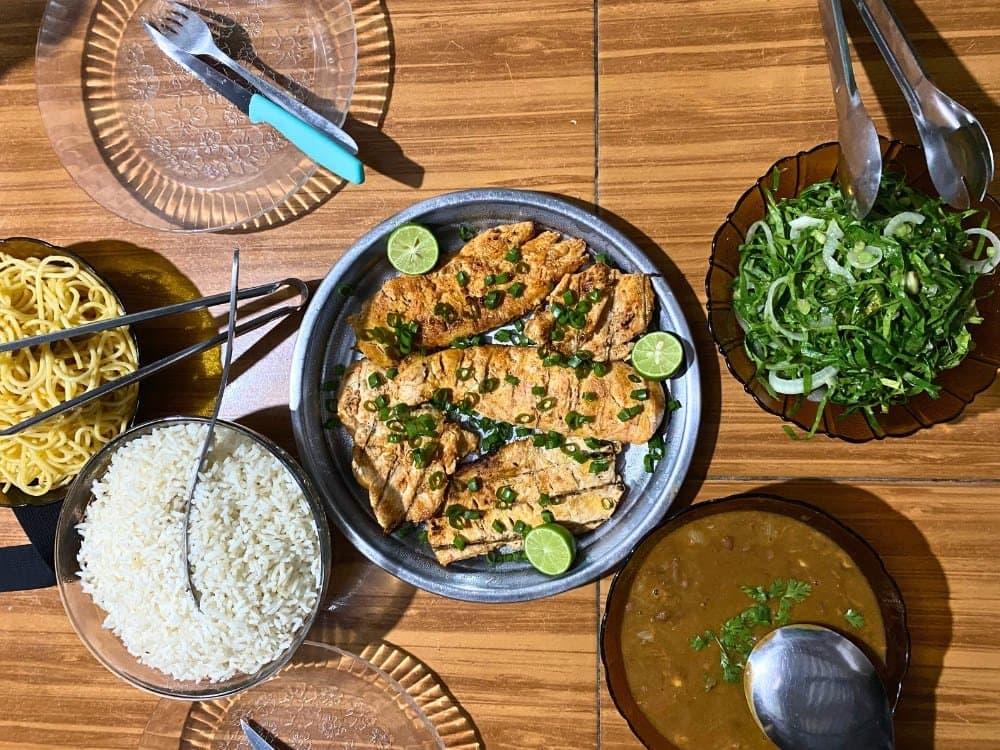 This is how you eat after a morning in the dunes. No fuss, all flavour. Overhead view of a home-cooked meal on a wooden table during a Lençóis Maranhenses trip, featuring grilled fish fillets topped with spring onion and lime, white rice, a green herb salad, bean stew, and yellow pasta noodles