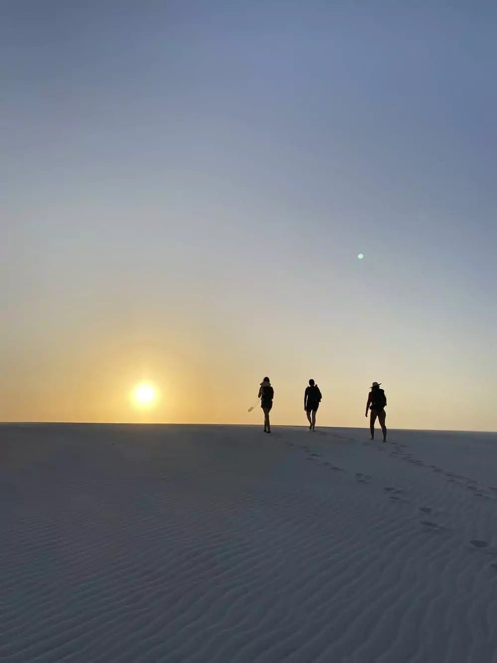 The dunes don't care about your schedule. The sun, however, is perfectly on time. Three travelers silhouetted against a golden sunset sky on a vast white sand dune during a Lençóis Maranhenses trip, footprints trailing behind them