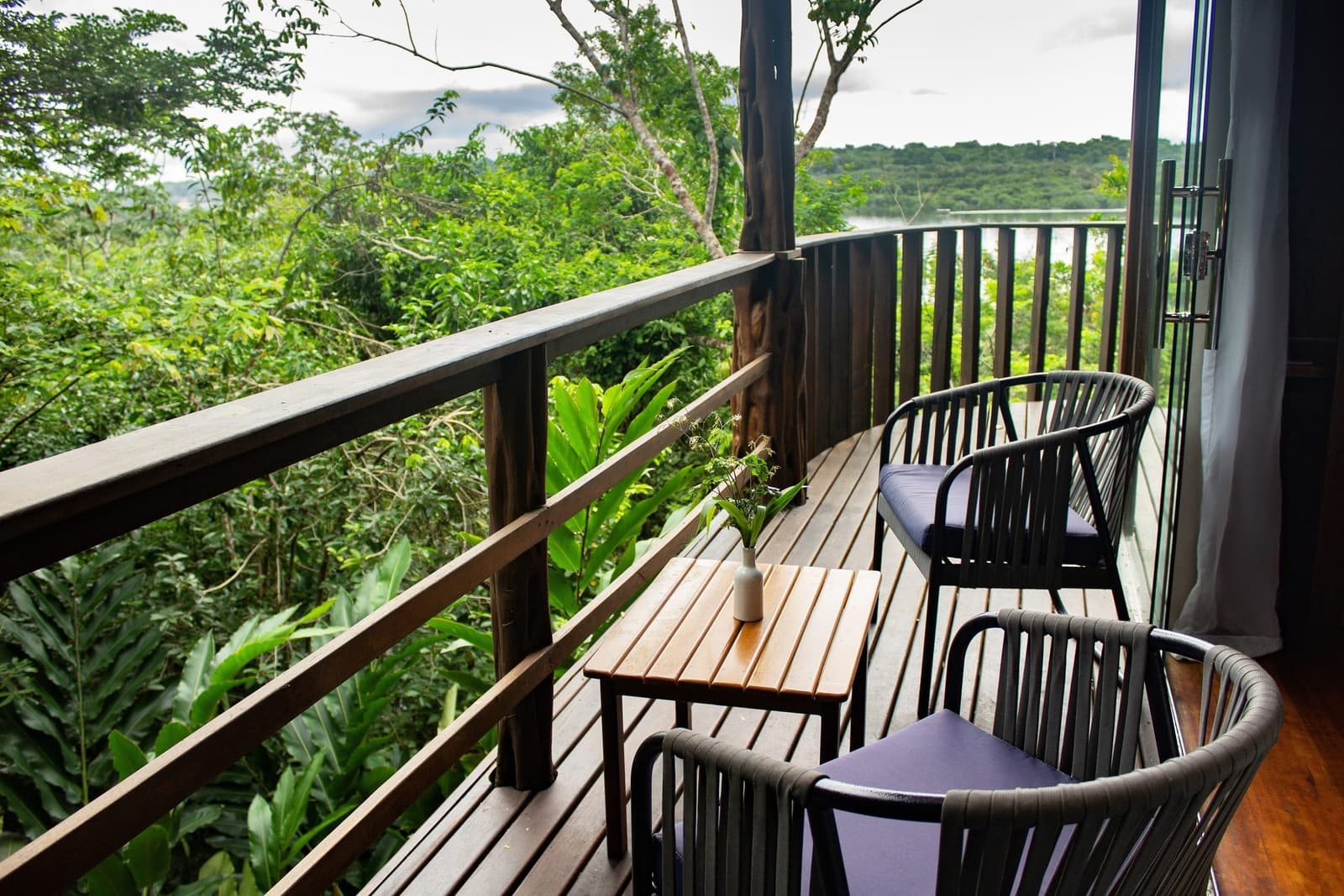 Your morning coffee has better scenery than most paintings. A curved wooden balcony at a luxury amazon lodge brazil with two rattan chairs and a small teak table overlooking dense green rainforest canopy and a distant river