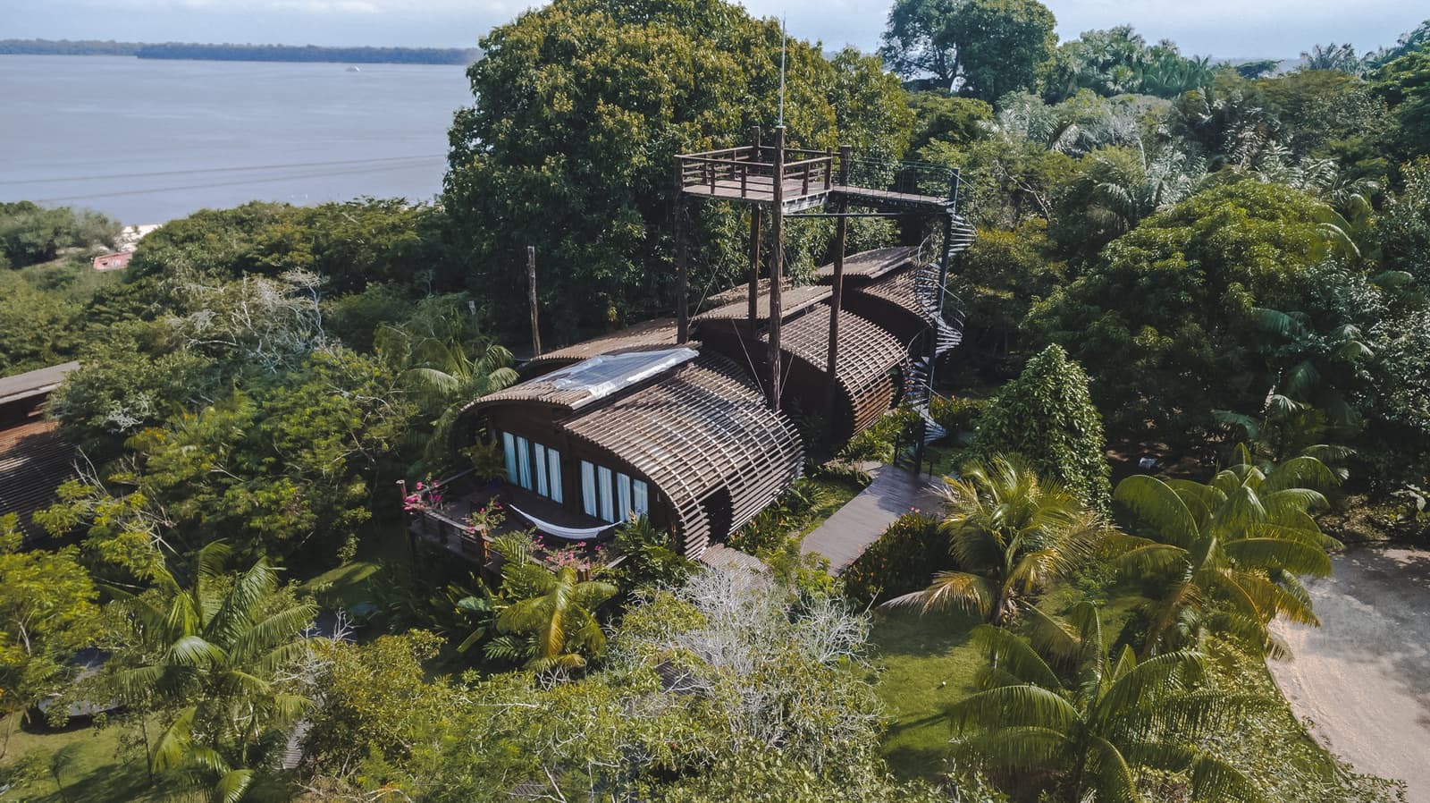 The canopy walkway on the roof tells you this place takes birdwatching seriously. Aerial view of a luxury Amazon lodge Brazil, showing a curved hardwood building with a slatted timber roof, a treetop canopy walkway rising above the rainforest canopy, and a wide river visible in the background