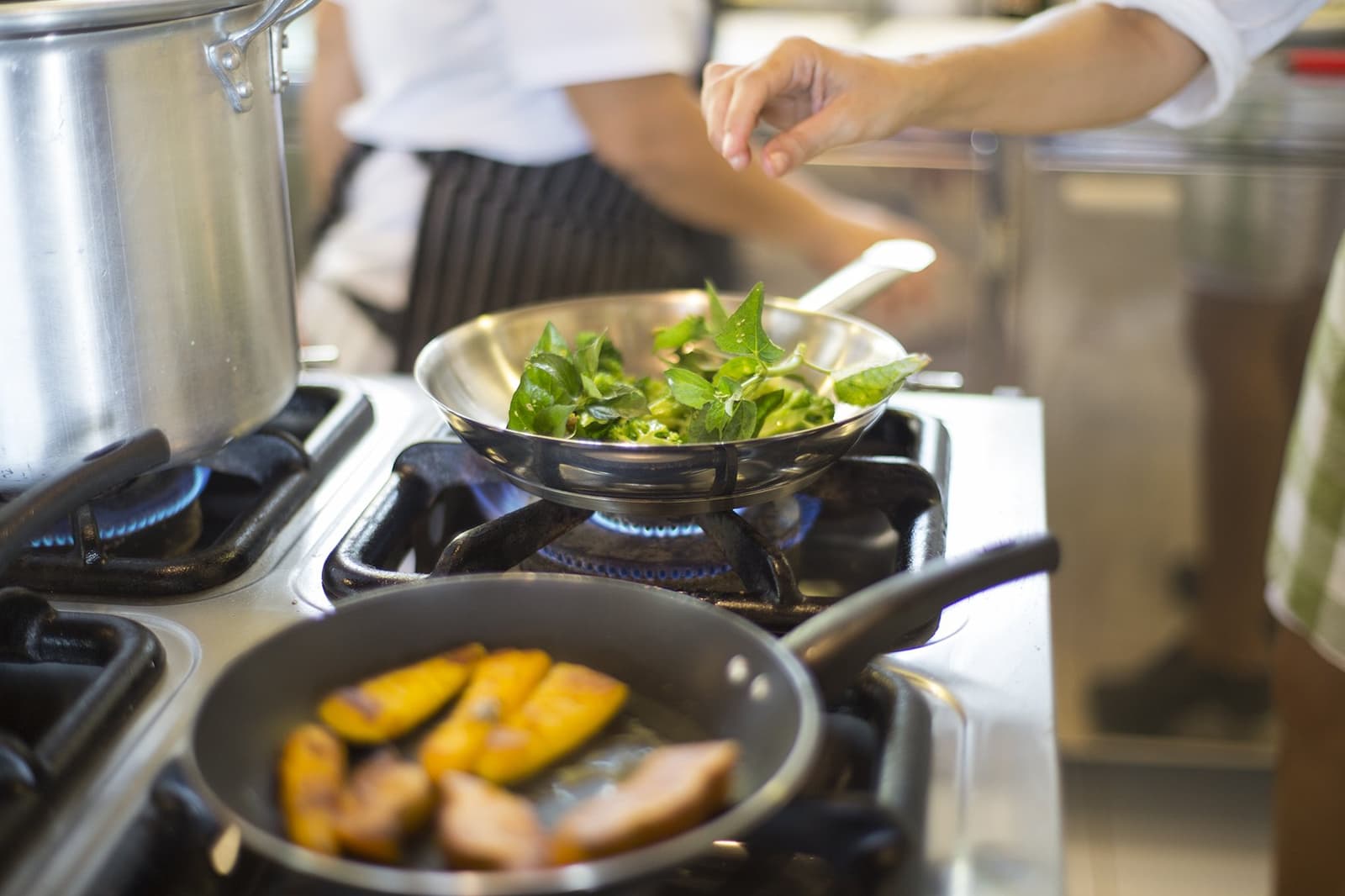 Local ingredients, live flame, and a cook who means business. A chef in a striped apron sautéing fresh green leaves in a stainless steel pan over a gas flame at a luxury Amazon lodge Brazil kitchen, with golden-seared fruit slices in a second pan in the foreground