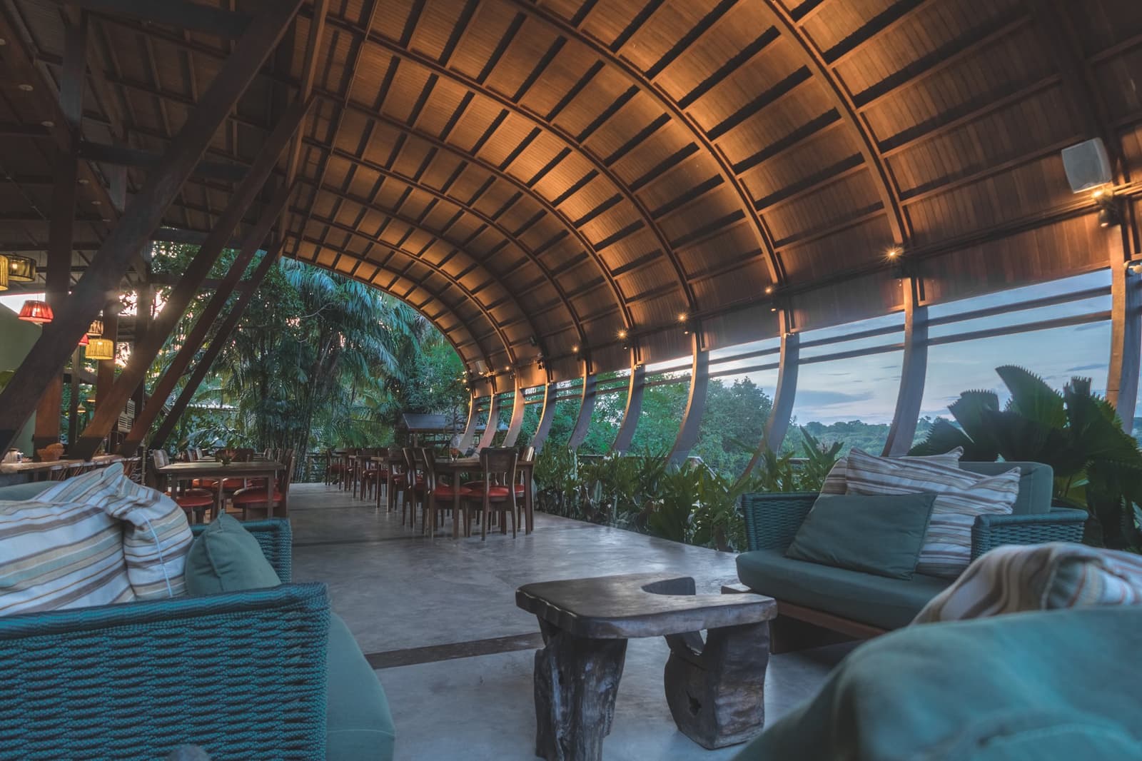 The kind of spot where you sit down for five minutes and lose an hour. Sweeping barrel-vaulted timber ceiling over the open-sided restaurant lounge of a luxury Amazon lodge in Brazil, with wicker sofas, a rustic log coffee table, and floor-to-ceiling views of lush rainforest canopy at dusk