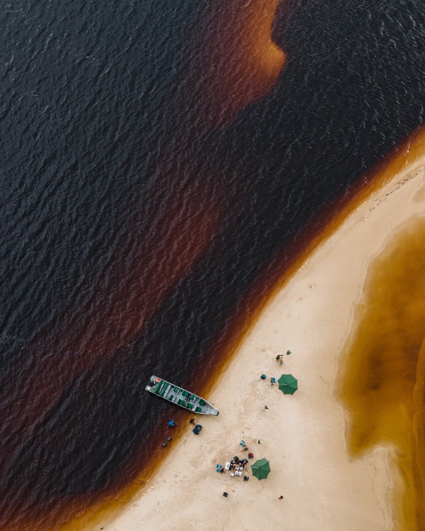 Two rivers, one sandbar, zero plans to leave anytime soon. Aerial view of a small motorboat beached on a white-sand bar beside the dark tannin-rich waters of a luxury Amazon lodge Brazil river excursion, with two green parasols and a group of guests gathered on the sand